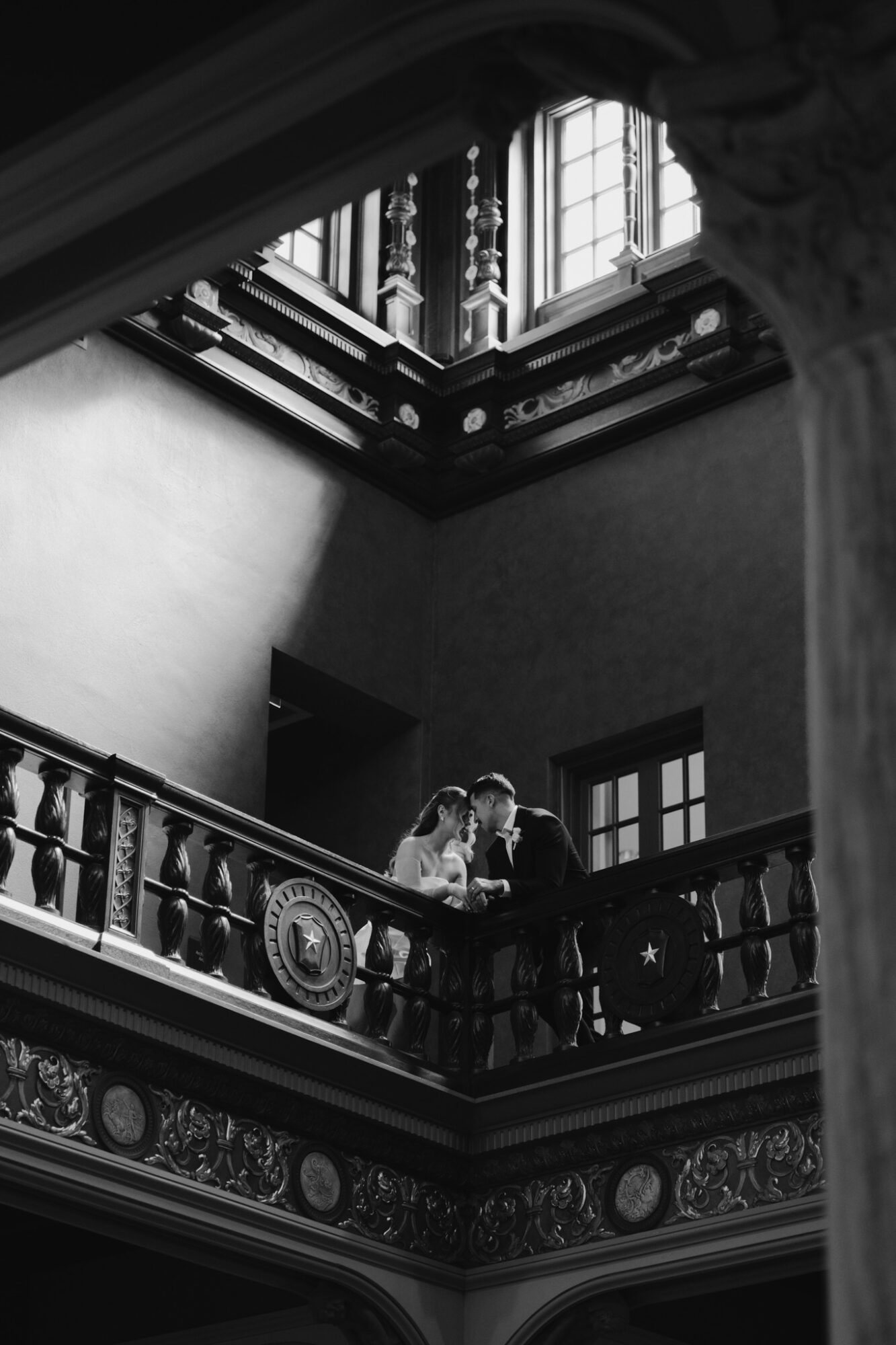 Two people stand on a balcony inside a grand, ornate building with large windows and detailed architectural features.