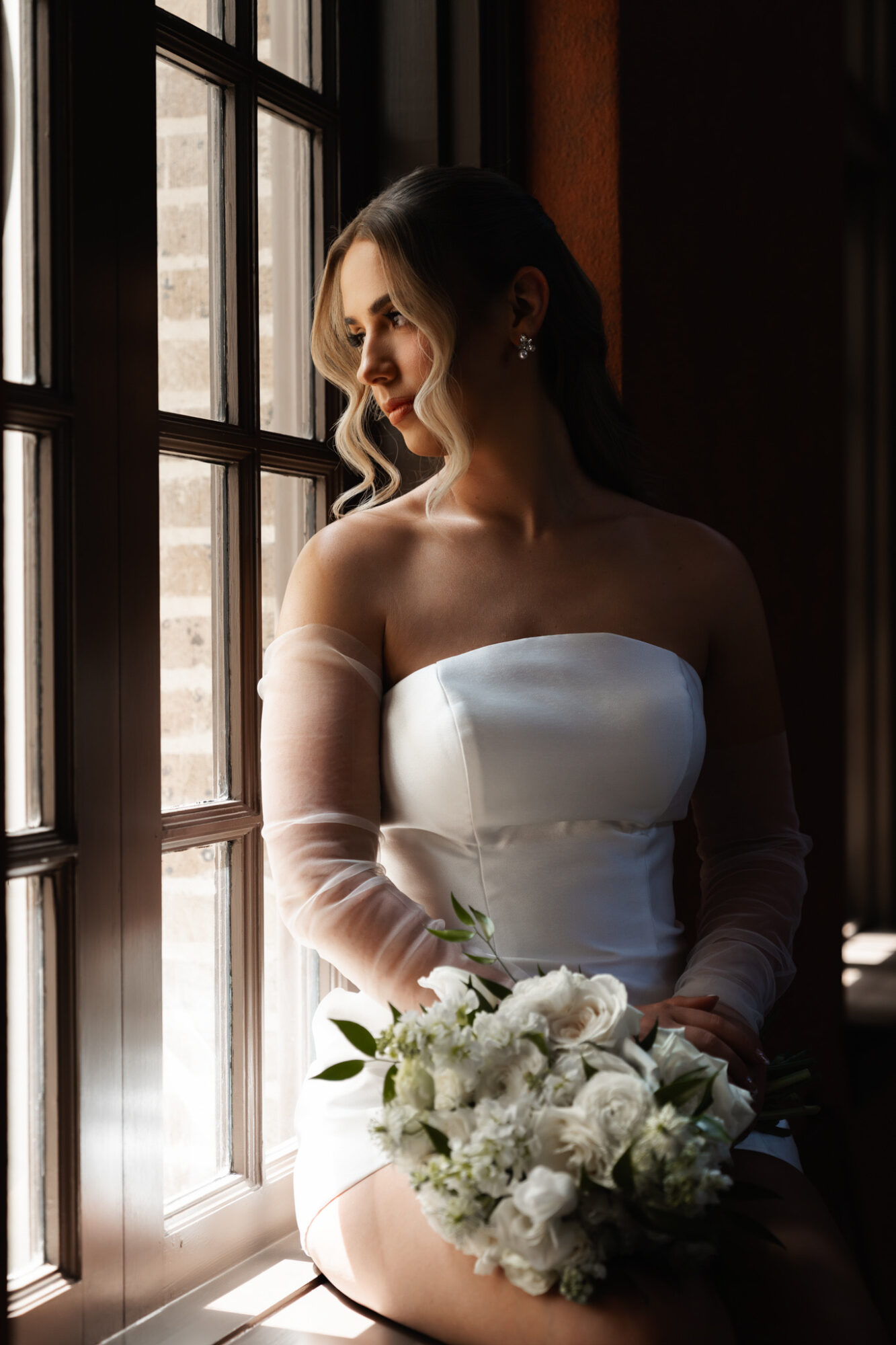 Woman in wedding dress holding bouquet, looking out window with sunlight illuminating her face.