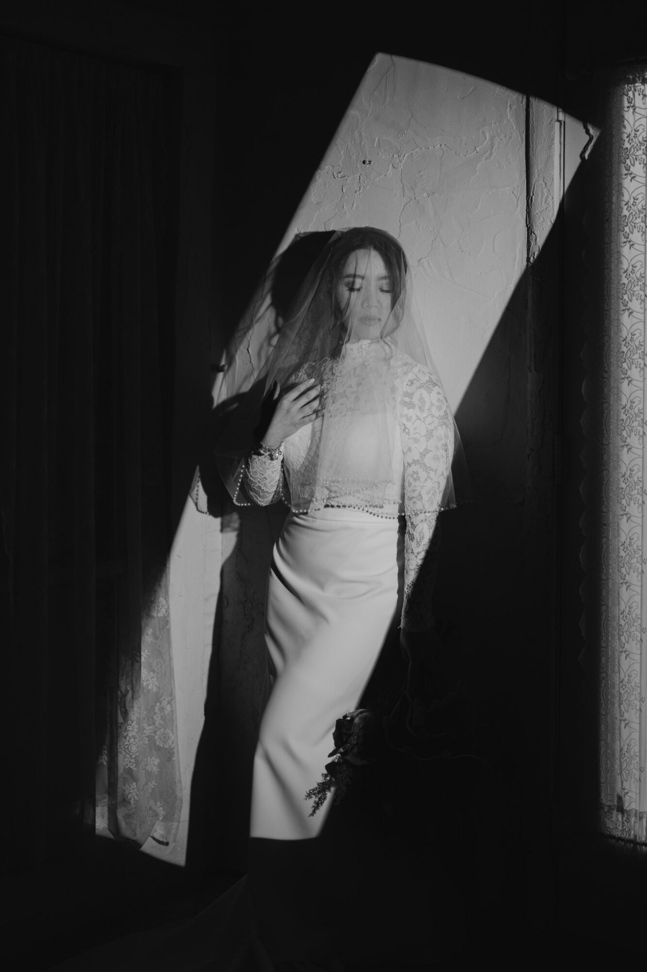 Woman standing in a doorway with light and shadow patterns, wearing a dress and veil, looking to the side.