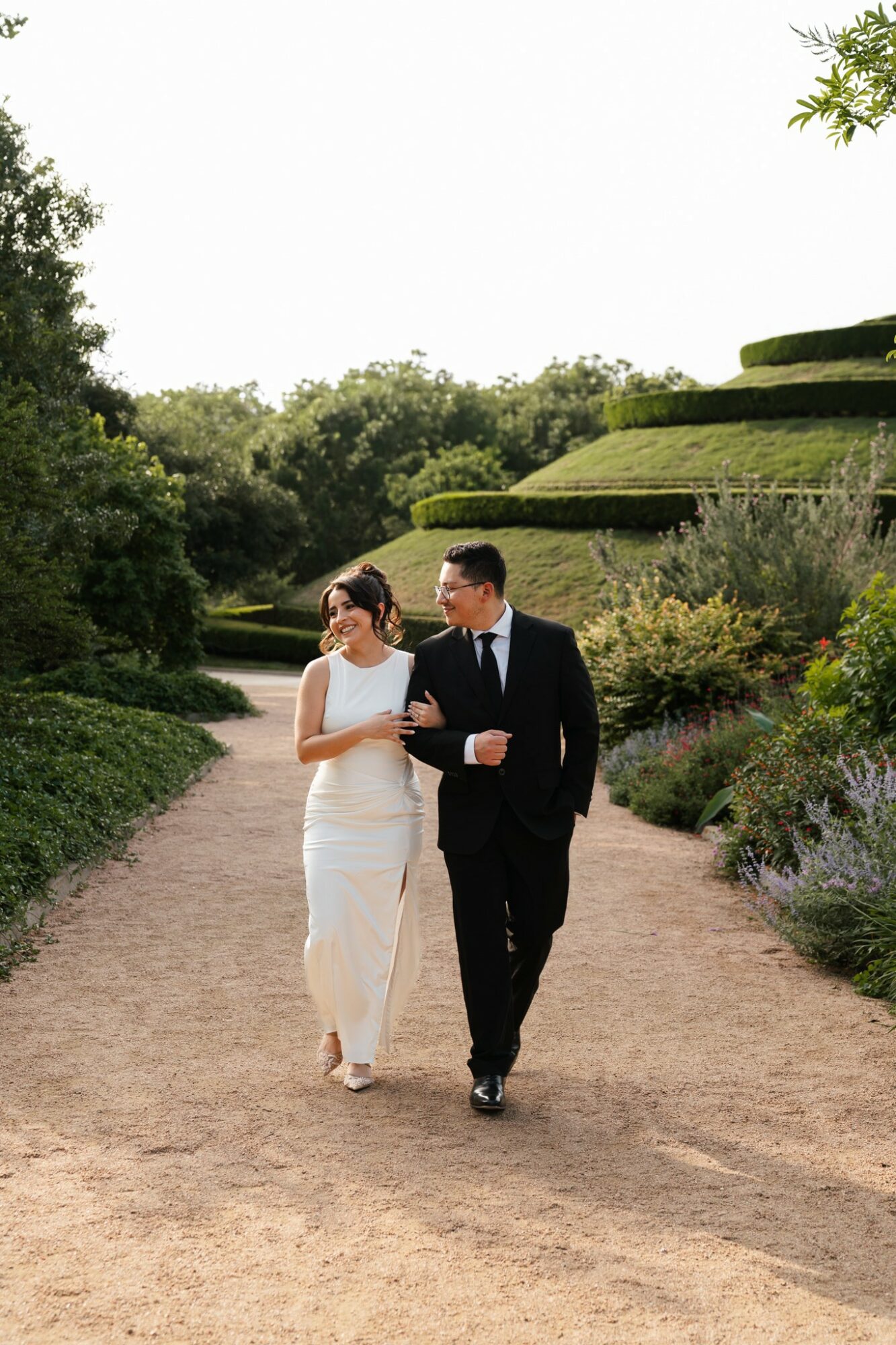 A couple in formal attire walking arm-in-arm on a garden path with tiered greenery in the background.