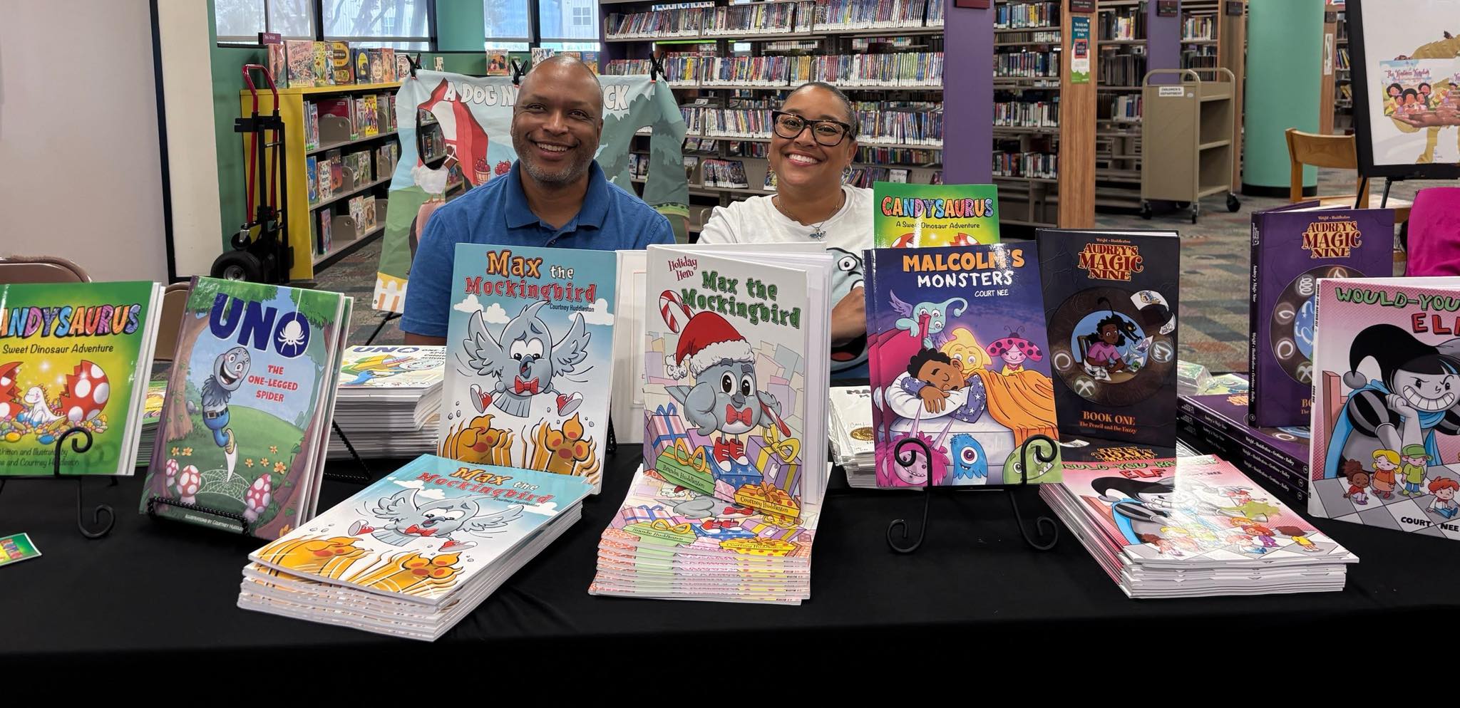 Two people sit behind a table with children's books in a library, smiling at the camera.