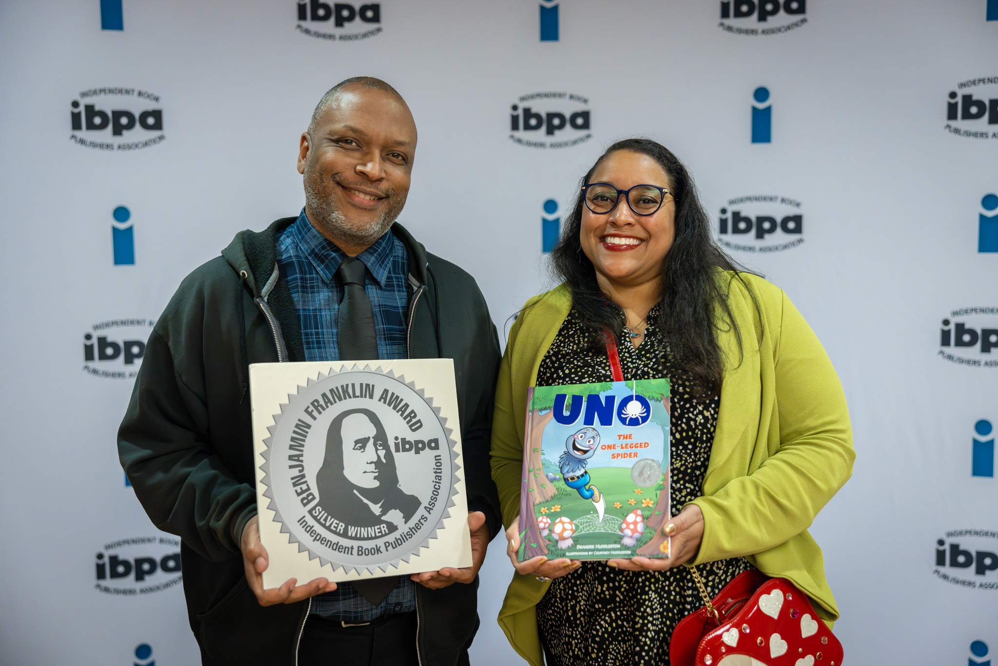 Two smiling people holding books in front of a backdrop with 'ibpa' logos, one man and one woman, both wearing glasses.