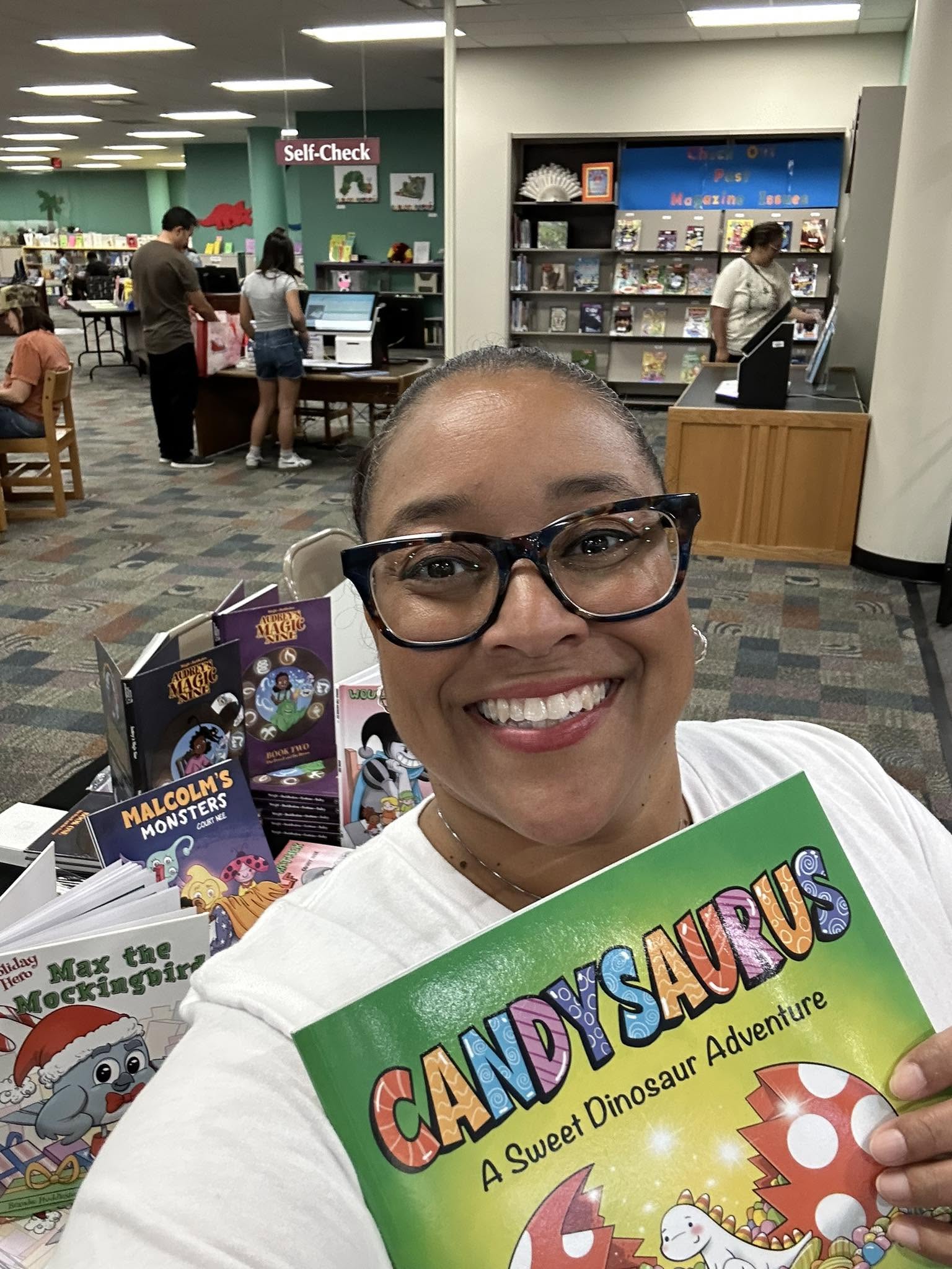 Smiling woman with glasses holding a Candy Safari book in a bookstore, with children and shelves in the background.