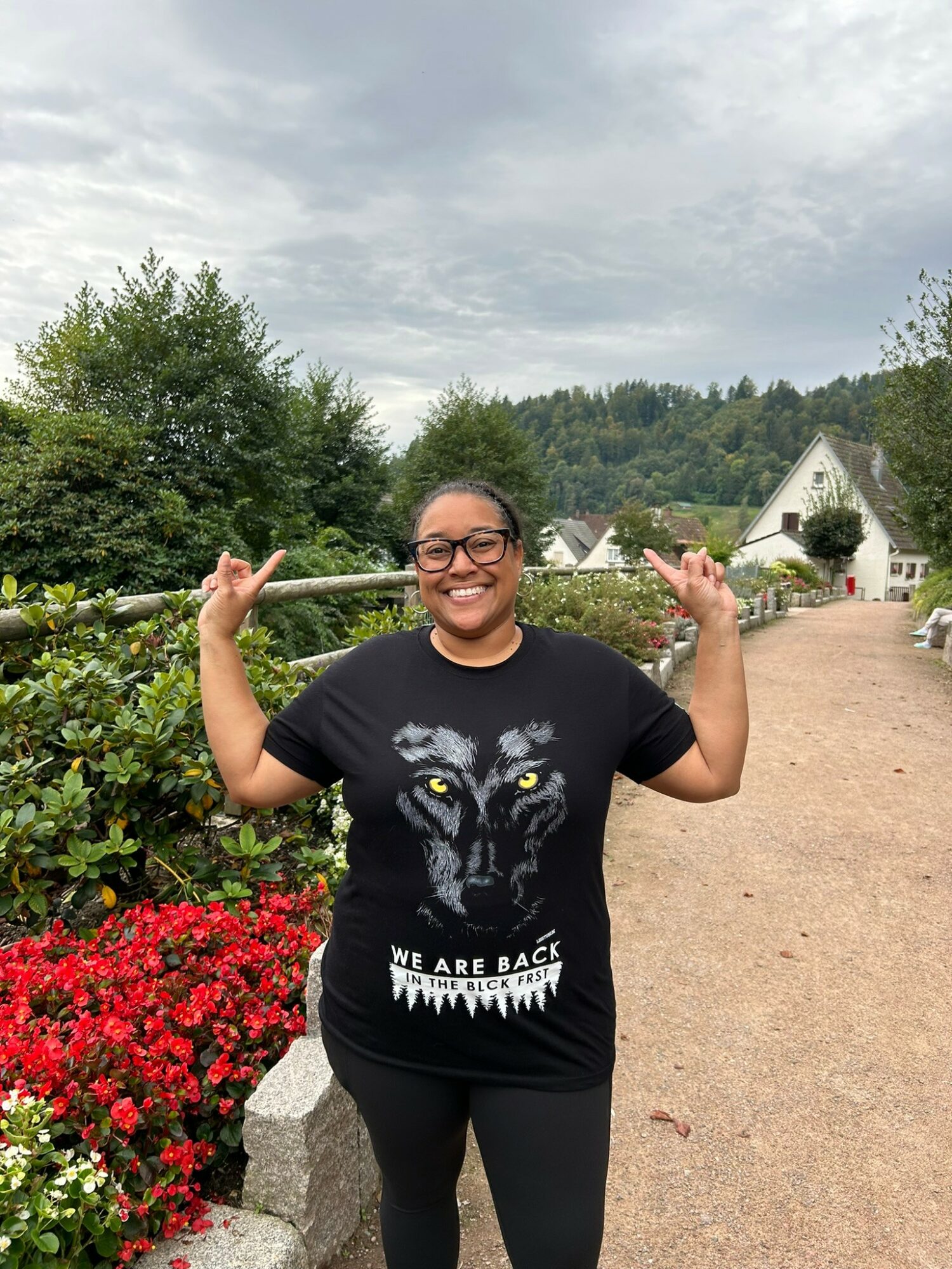 Smiling woman with glasses and a black t-shirt standing outdoors, holding a stick over her shoulders, surrounded by greenery and flowers.