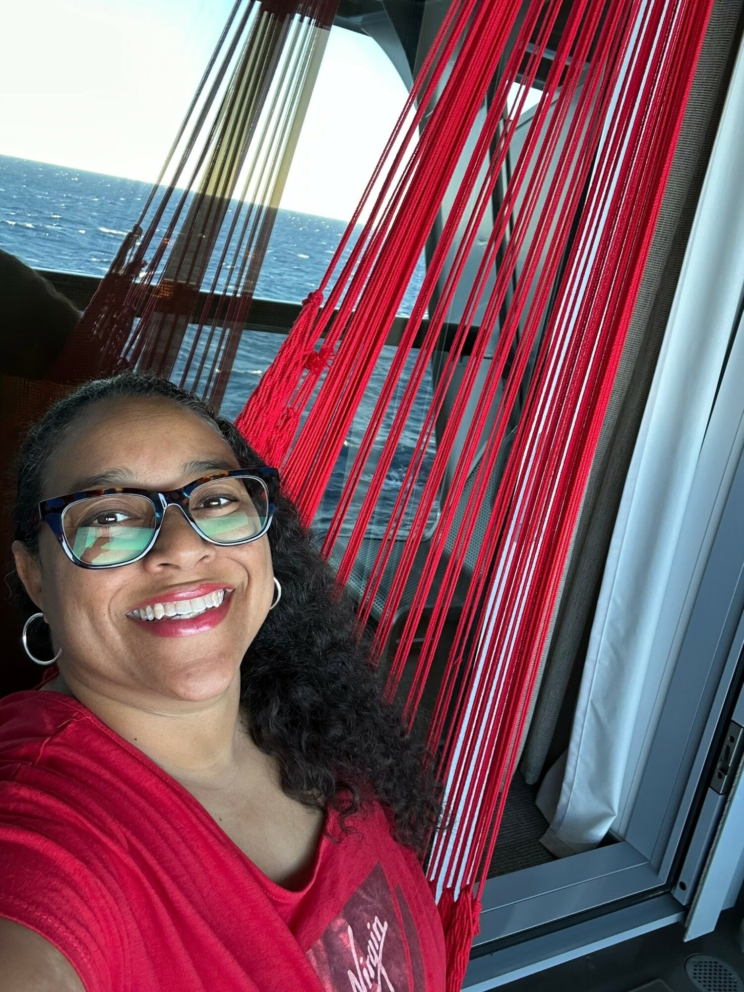 Smiling woman with glasses and curly hair in front of a window with red and black curtains, ocean view in background.