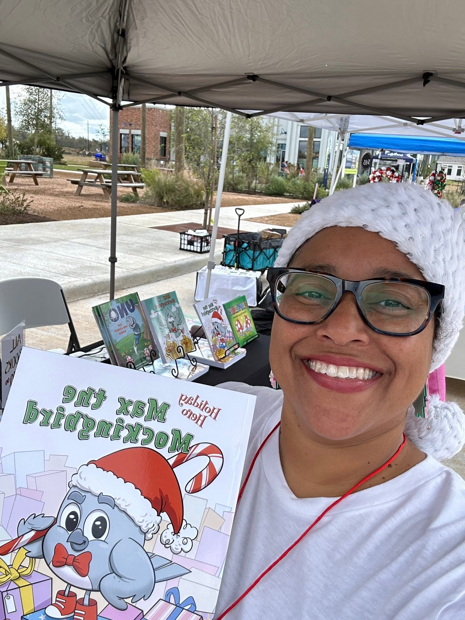 Smiling woman wearing glasses and a Santa hat, holding a children's book with an elephant on the cover, outdoors under a canopy.