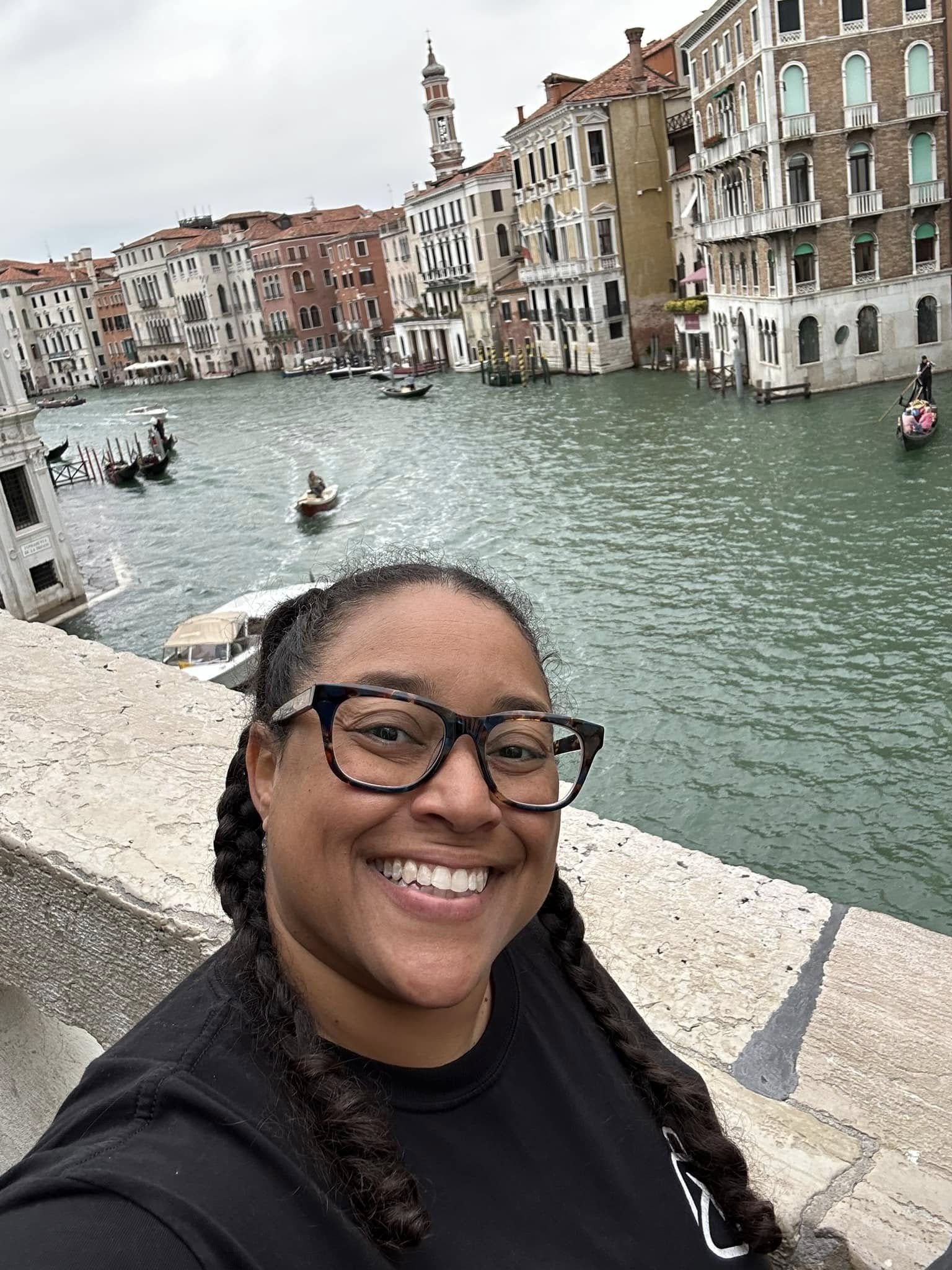 Smiling woman with glasses takes selfie with canal and historic buildings in Venice, Italy in the background.