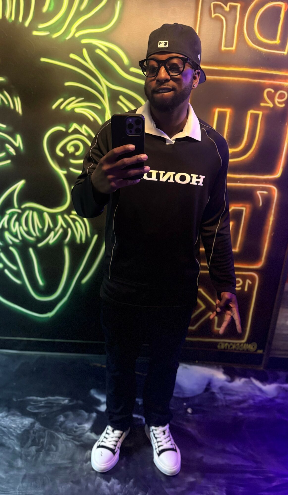 Person taking a selfie in front of neon art, wearing a black hoodie, cap, glasses, and white sneakers.