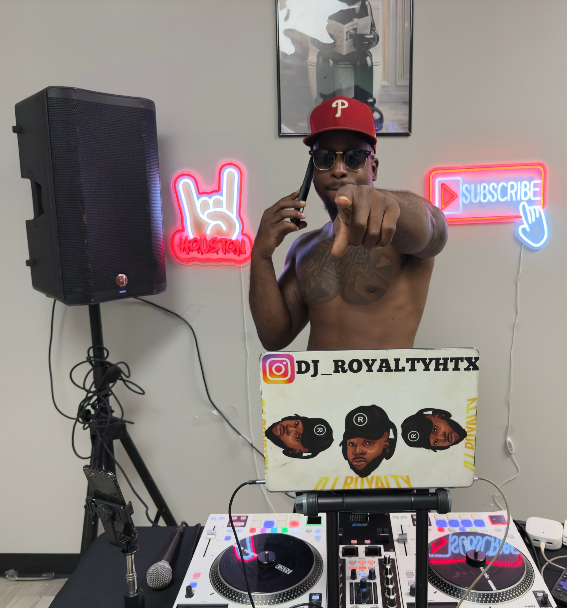 Shirtless man wearing sunglasses and a red cap, pointing at the camera, with DJ equipment and neon signs in background.