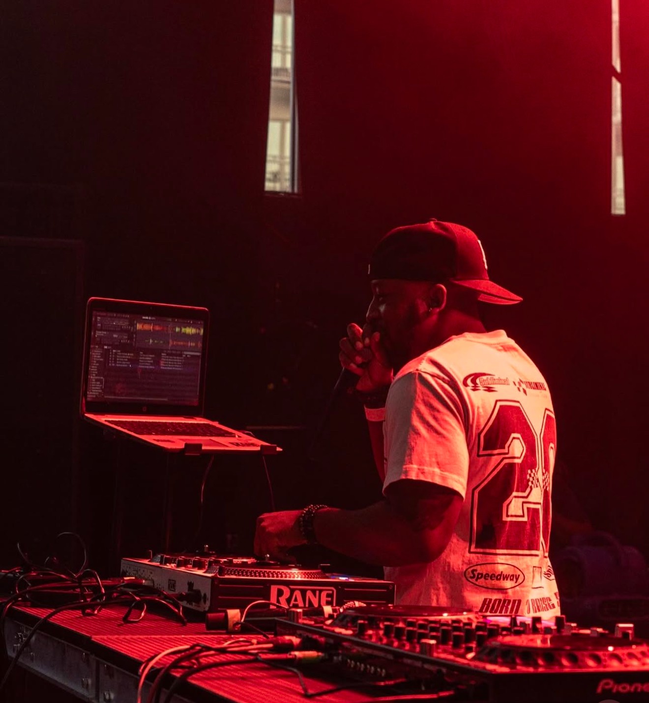 Person DJing with laptop and equipment, wearing a cap and sports jersey, in a dark setting with red lighting.