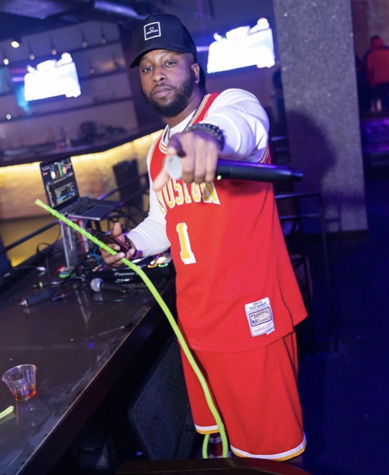 Man in red basketball jersey and cap pointing at camera in a nightclub with DJ equipment behind him.