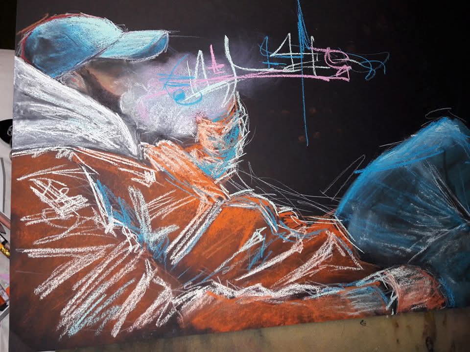 Person lying on the ground wearing a cap, jacket, and jeans, with colorful chalk drawings on a black surface.
