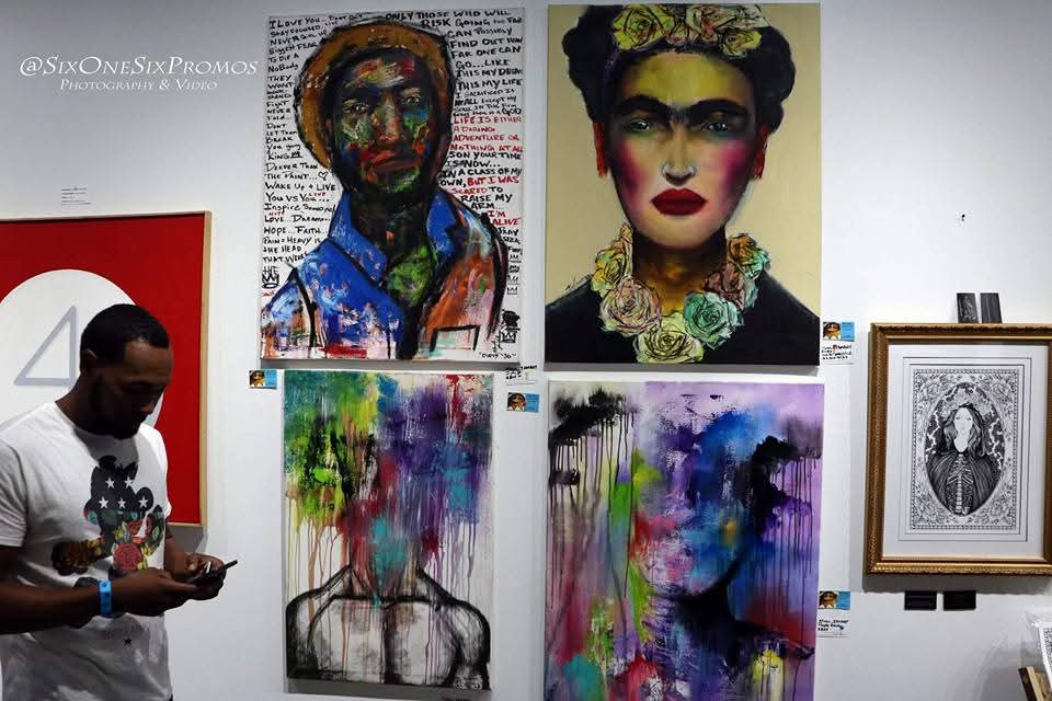 Gallery wall with colorful portraits, abstract paintings, and framed artwork, including a woman looking at her phone.
