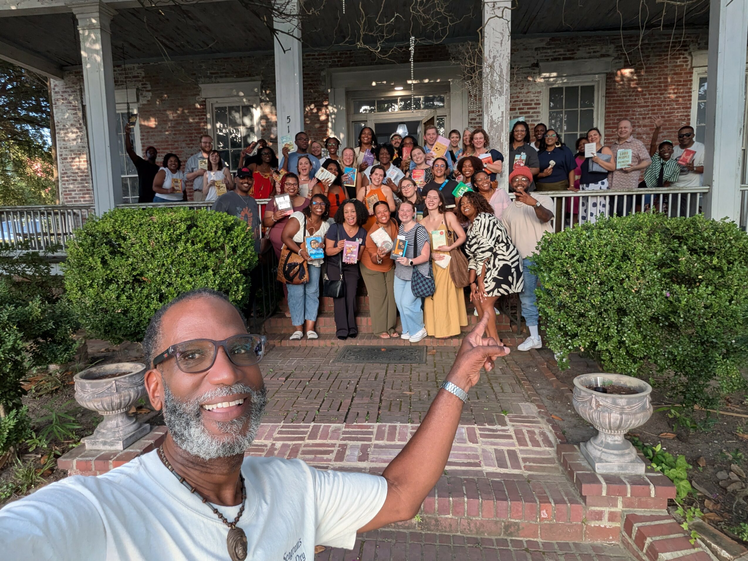 Group of people gathered on porch, with man in foreground taking selfie, smiling, pointing at group, greenery around, brick walkway.