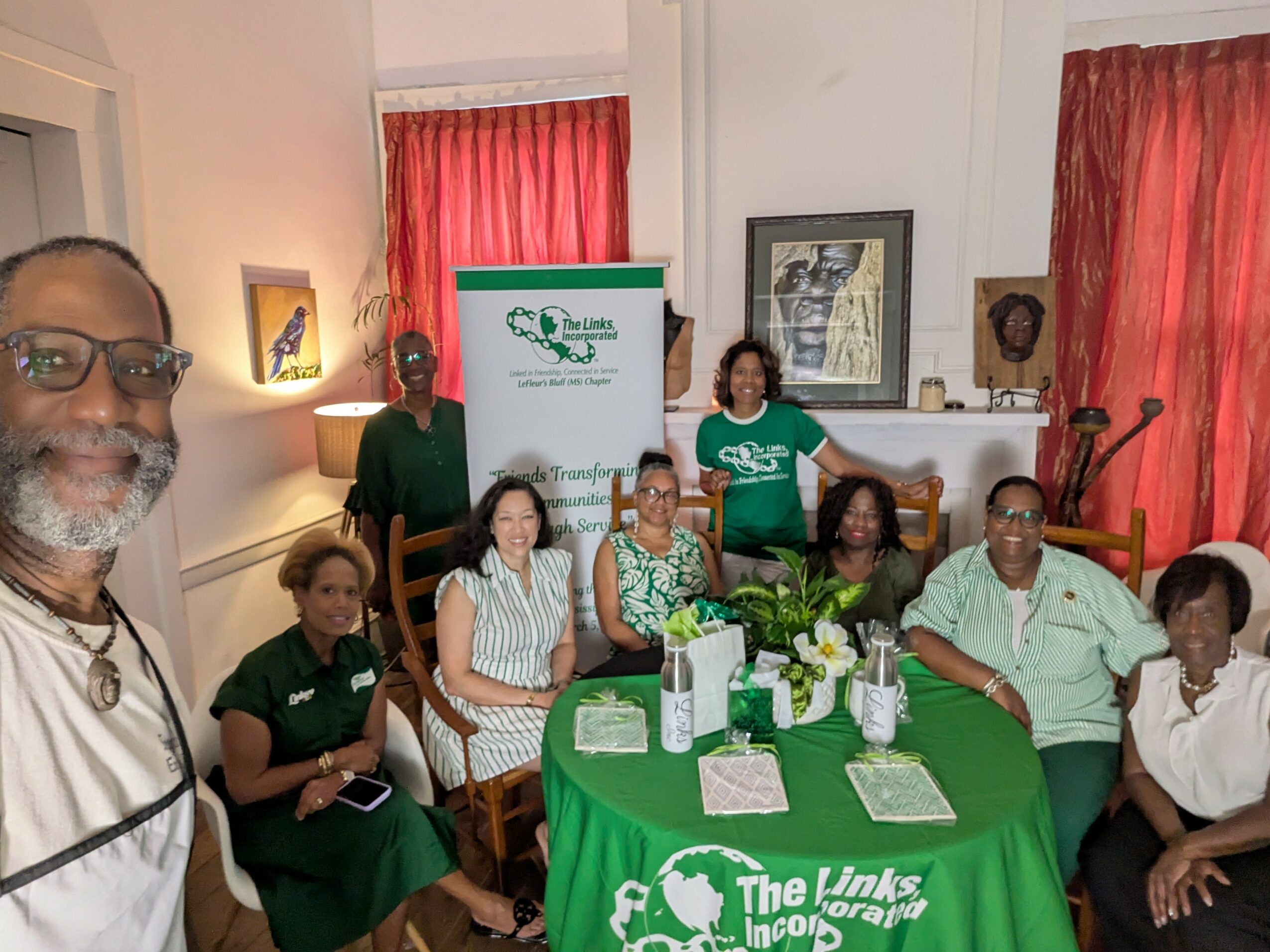Group of eight people sitting and standing around a table with green tablecloth, in a room with red curtains and artwork.