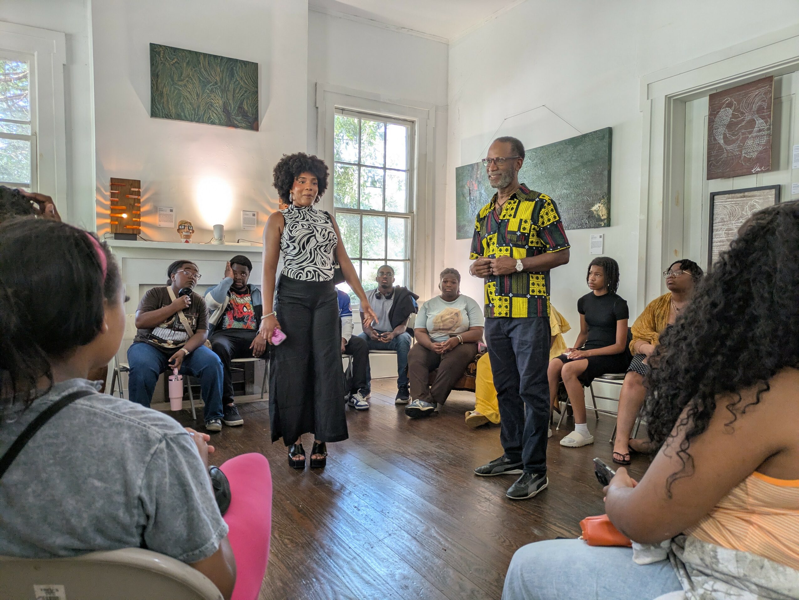 People gathered in a room with two individuals standing and speaking, others seated around in a circle, bright natural light.