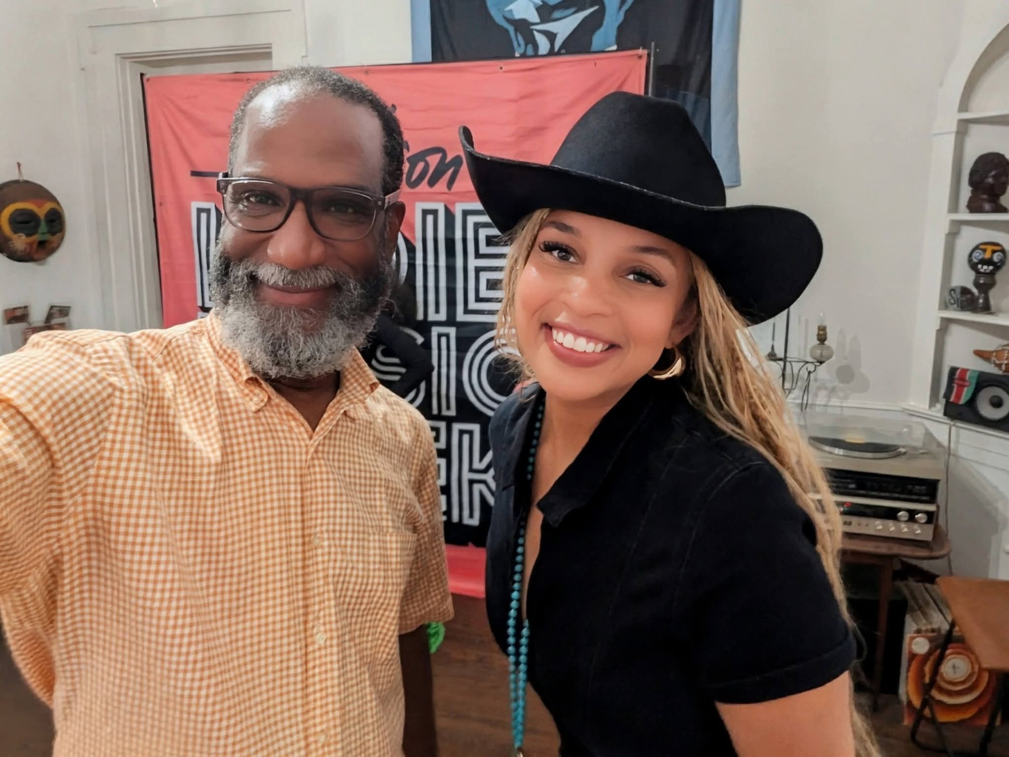 A smiling man with glasses and a beard wearing a checkered shirt, and a woman with a black hat and long hair, smiling, in an indoor setting.