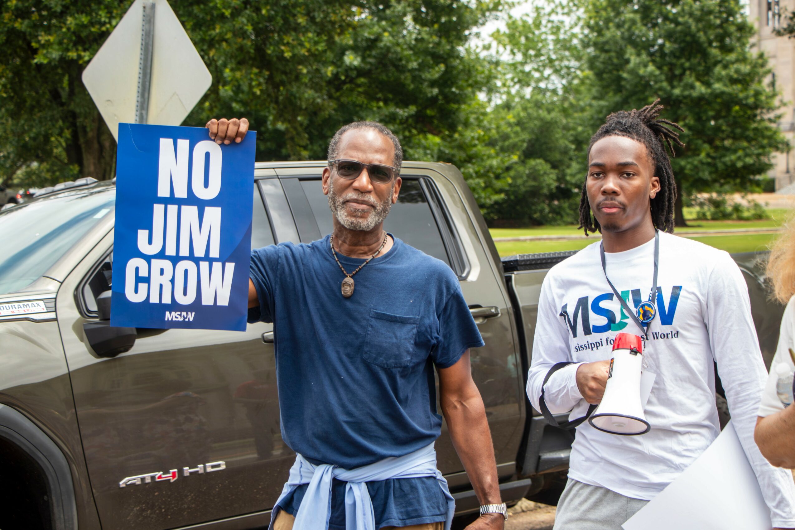Man holding a blue sign that reads 'NO JIM CROW' standing next to a woman with a megaphone outdoors.