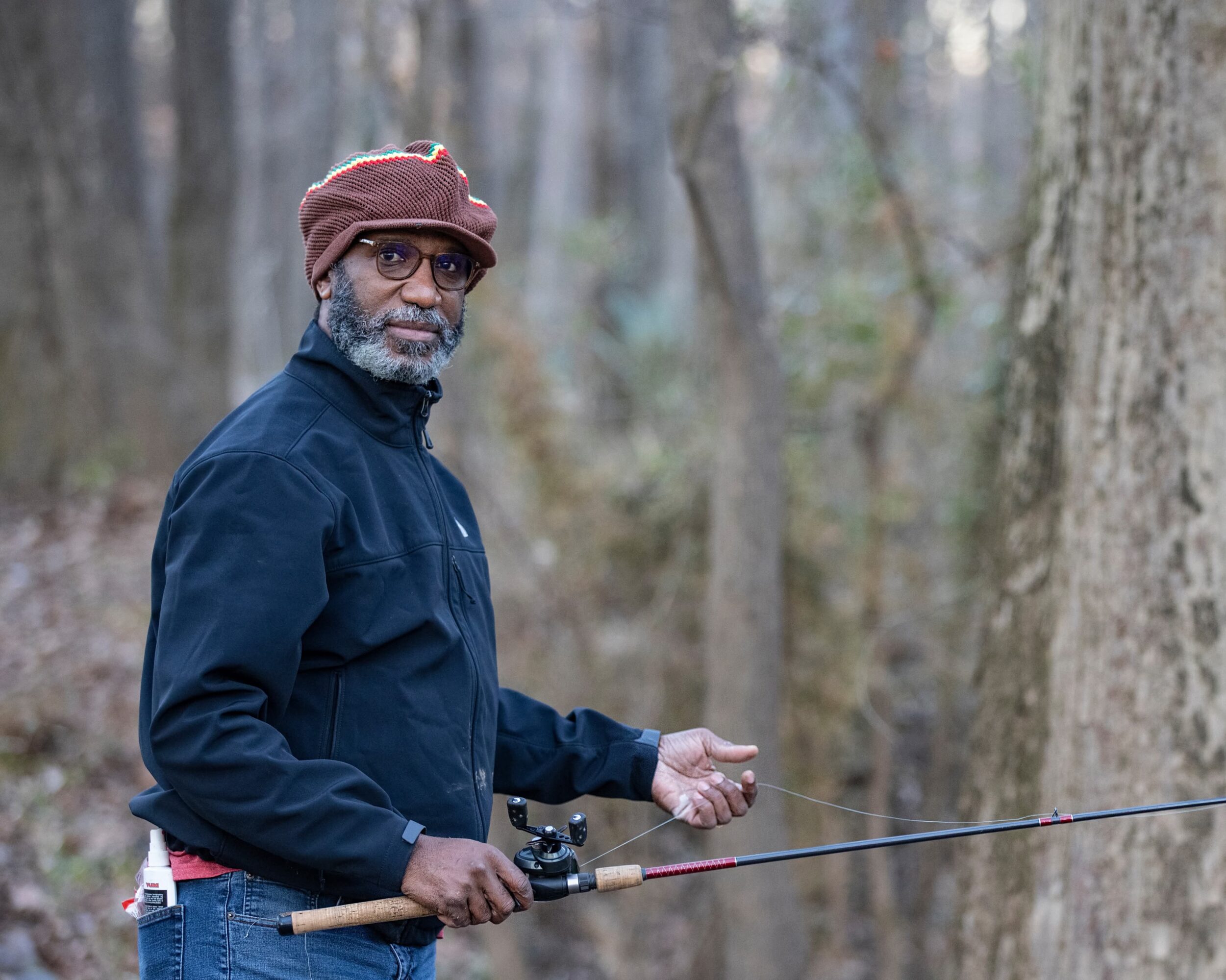 Man with glasses and a hat holding a fishing rod in a forest, looking at the camera.
