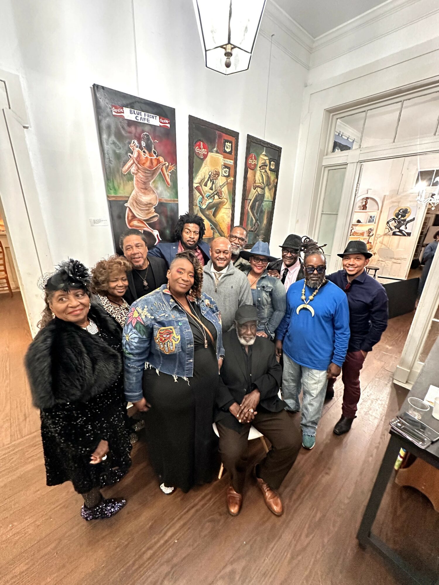 Group of people posing indoors in front of three posters, some wearing hats and glasses, smiling at the camera.