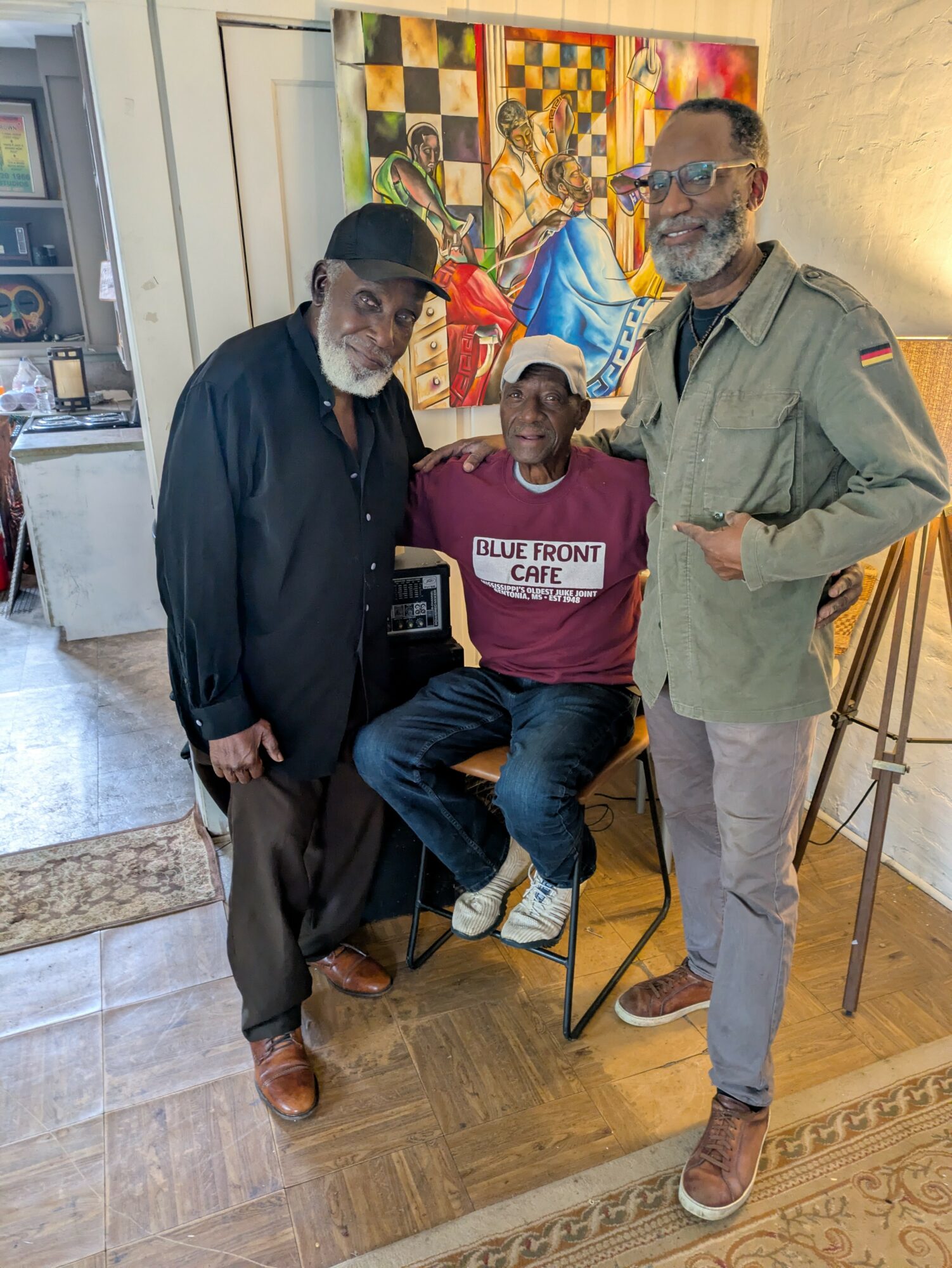 Three men pose indoors with colorful artwork in the background. One seated, two standing, all smiling. The seated man wears a maroon shirt, the others are in dark and light-colored clothing.