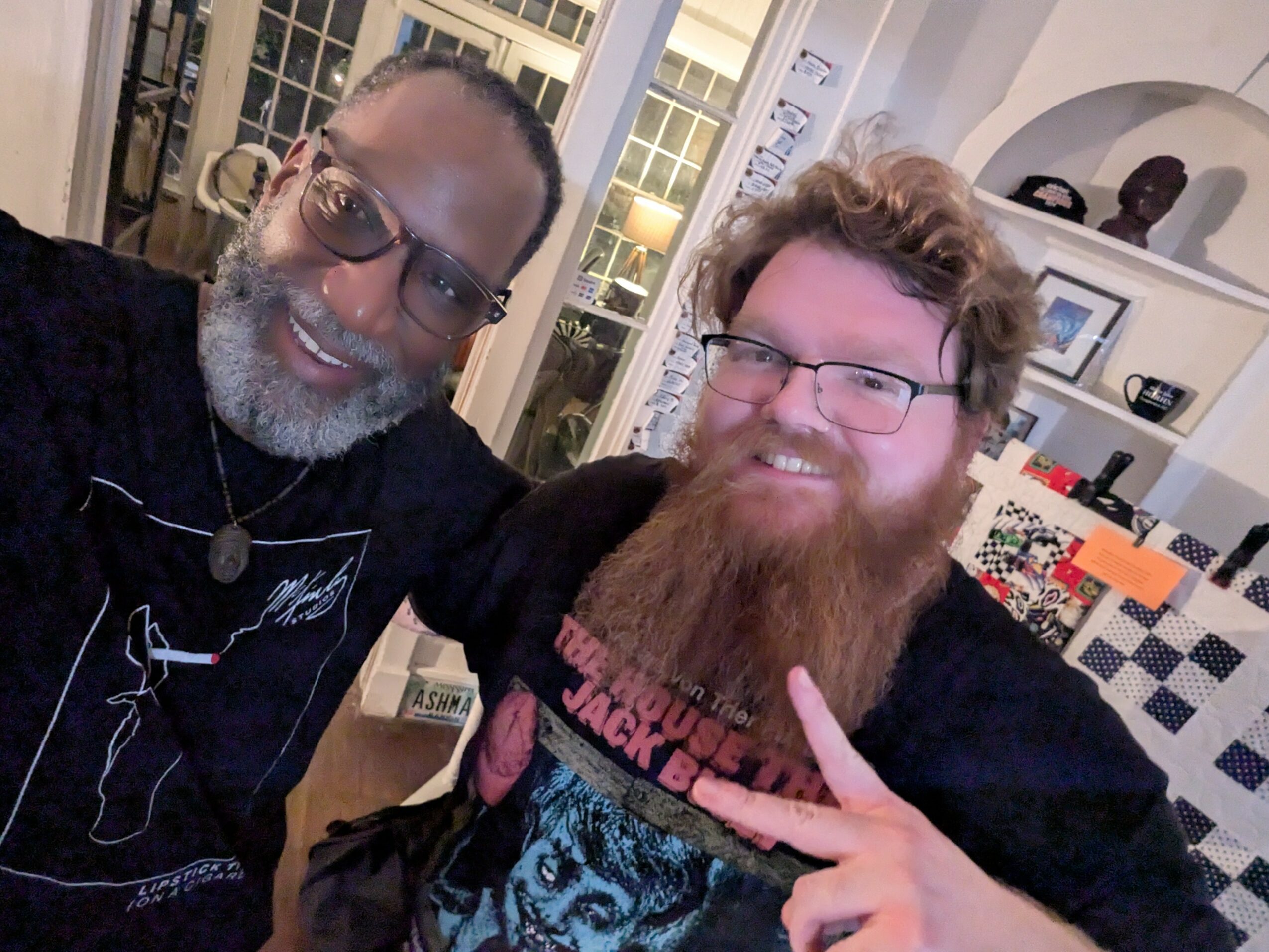 Two men with glasses and beards smiling indoors, one making a peace sign, background shows a kitchen and window.