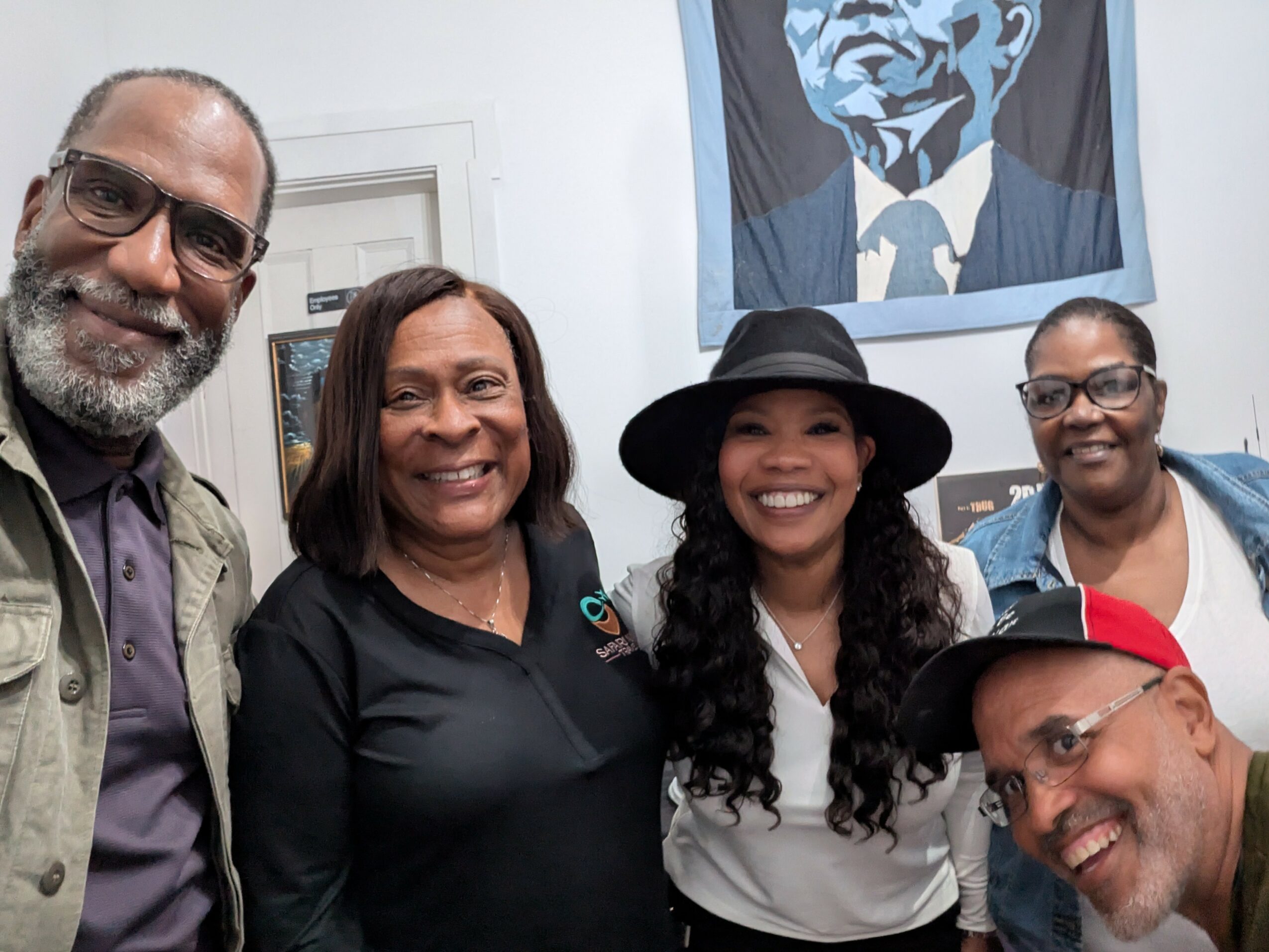 Four people smiling indoors, with a portrait of a man hanging on the wall behind them.