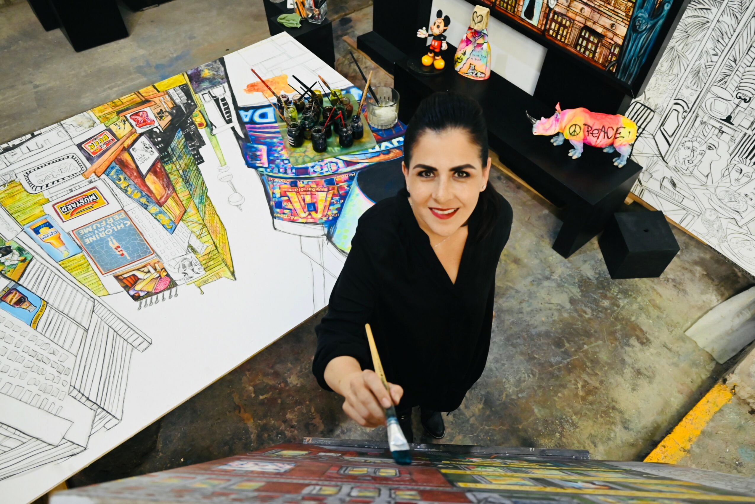 Woman with dark hair and black clothing holding a paintbrush, standing in an art studio with drawings and models around her.