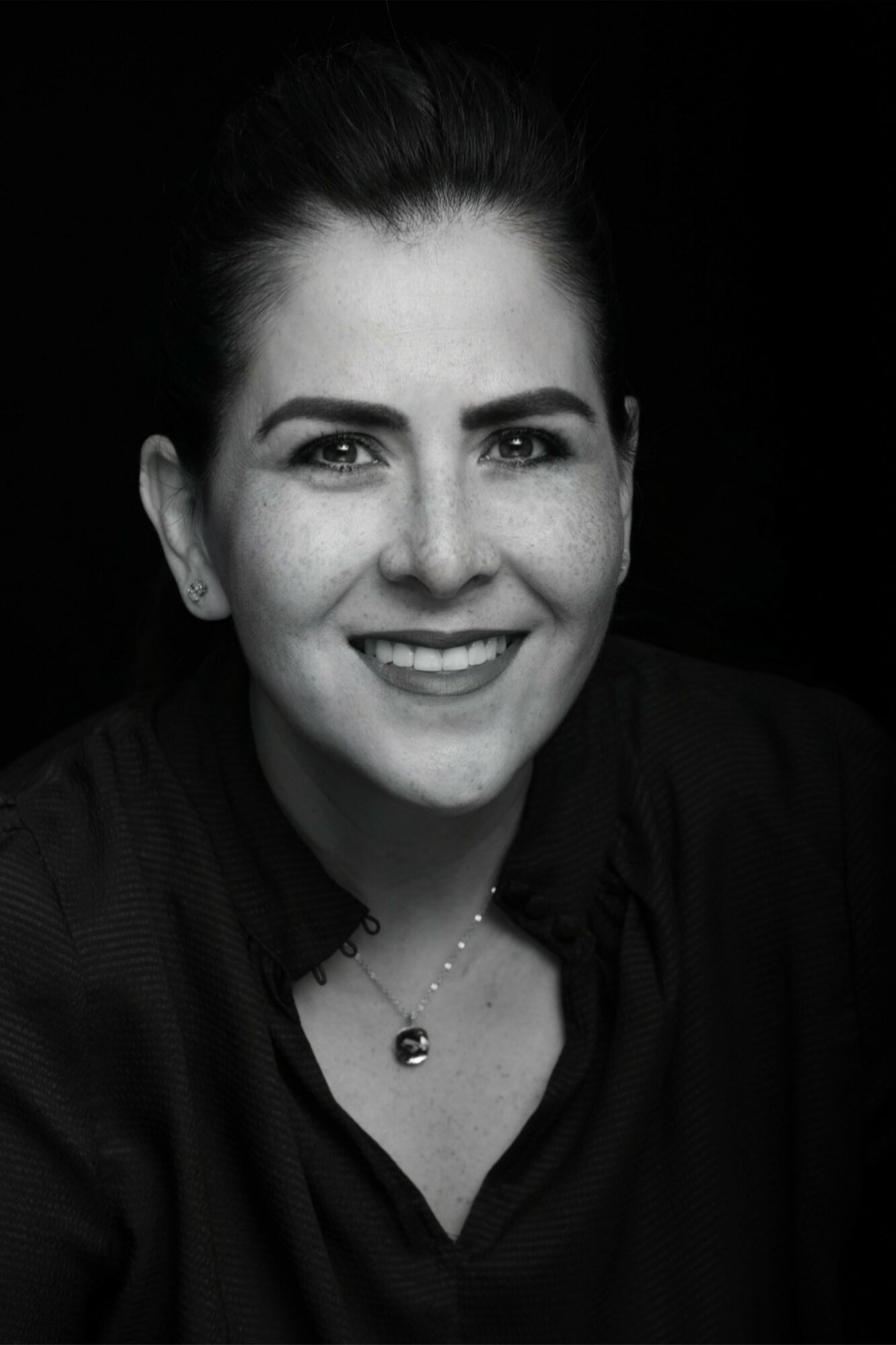 Portrait of a smiling woman with dark hair pulled back, wearing a dark top and a necklace, against a black background.