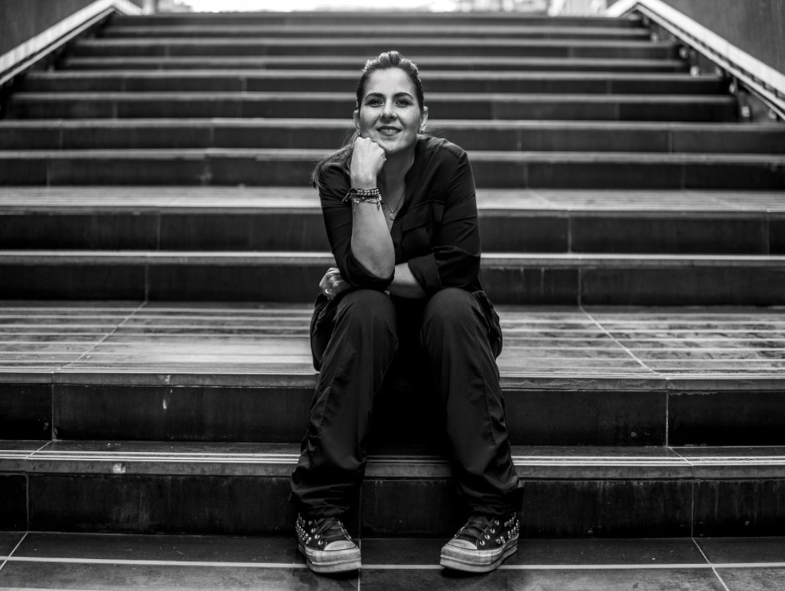 Woman sitting on stairs, smiling, resting her chin on her hand, wearing dark clothing and sneakers, black and white photo.