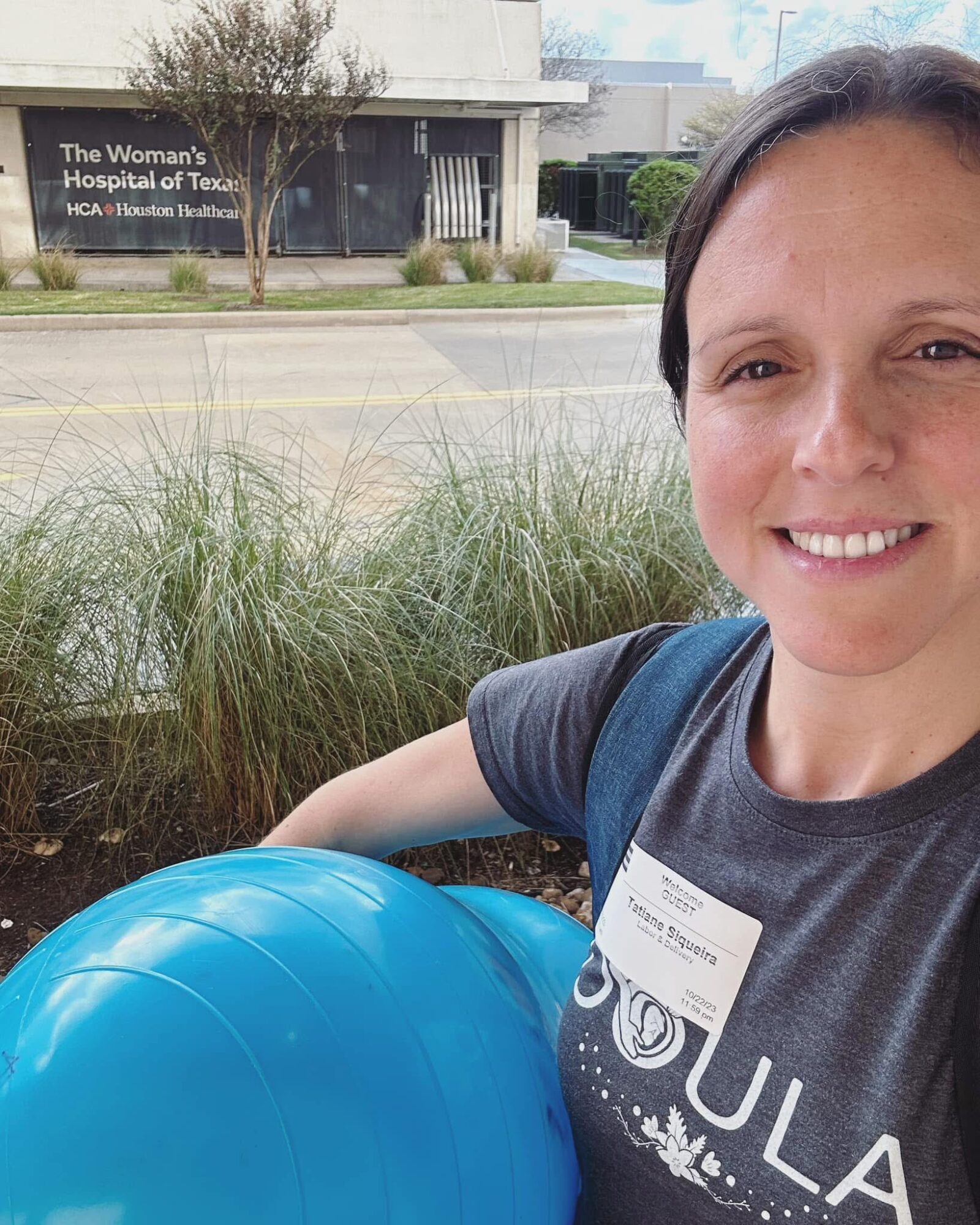 Smiling woman outdoors holding a blue exercise ball, with a hospital sign in the background.
