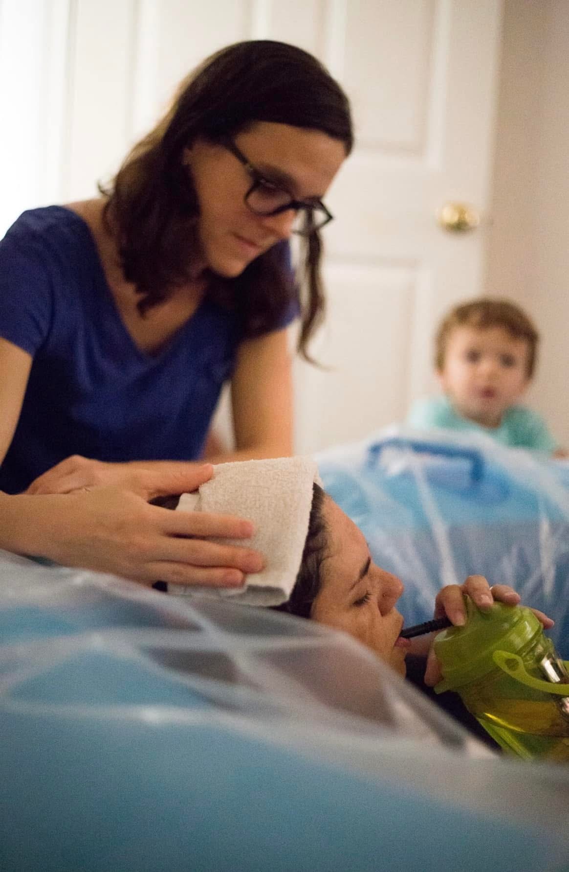 Woman with glasses holding a child lying down, with a young boy in the background, in a room with a door.