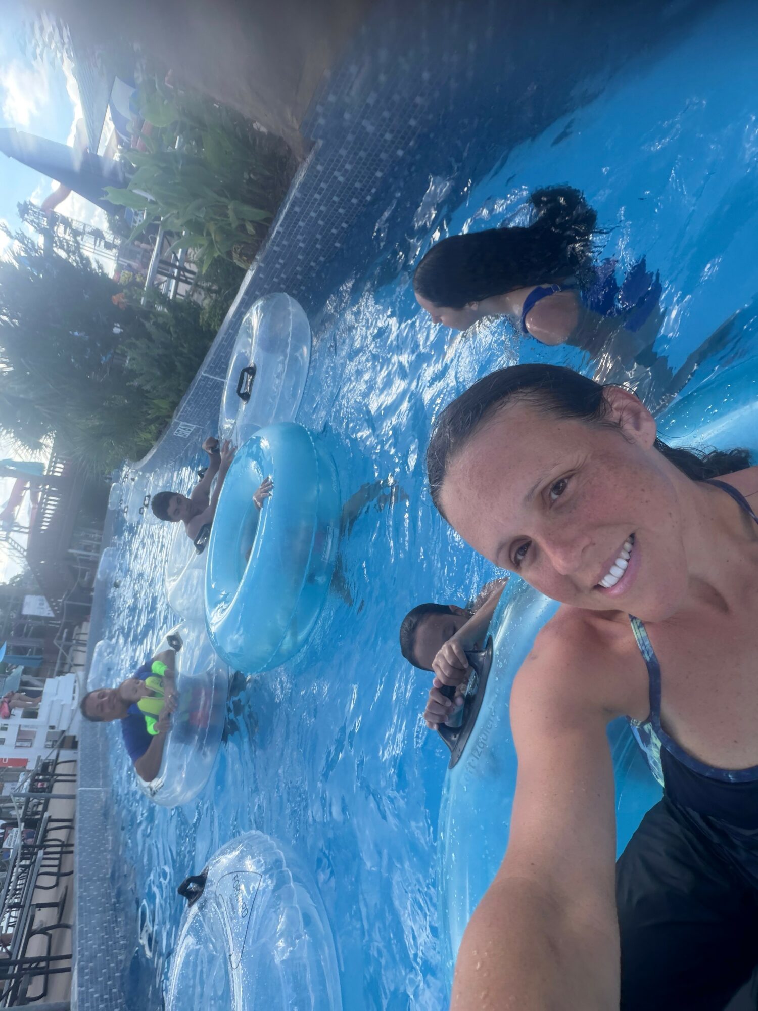 Woman smiling in a swimming pool with children, floating on inflatable rings, surrounded by water and trees.