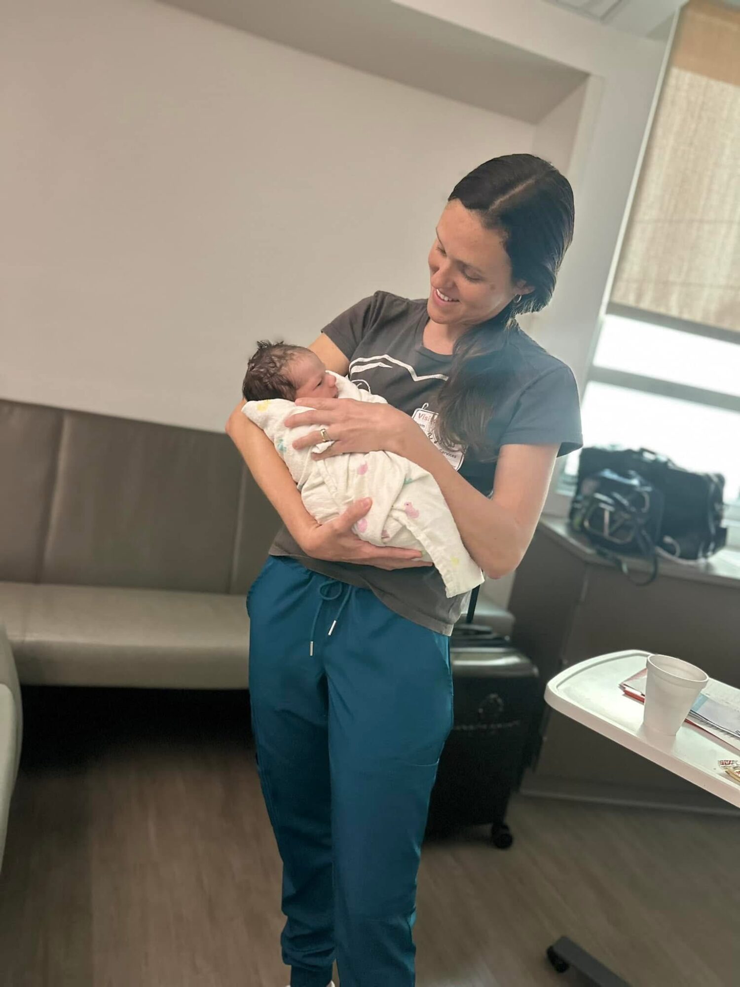 Woman holding a newborn baby in a room with a bench, window, and table with a cup.