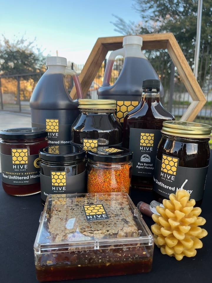 Assorted honey jars and bottles with honeycomb and a honey bee-shaped candle on a table outdoors.