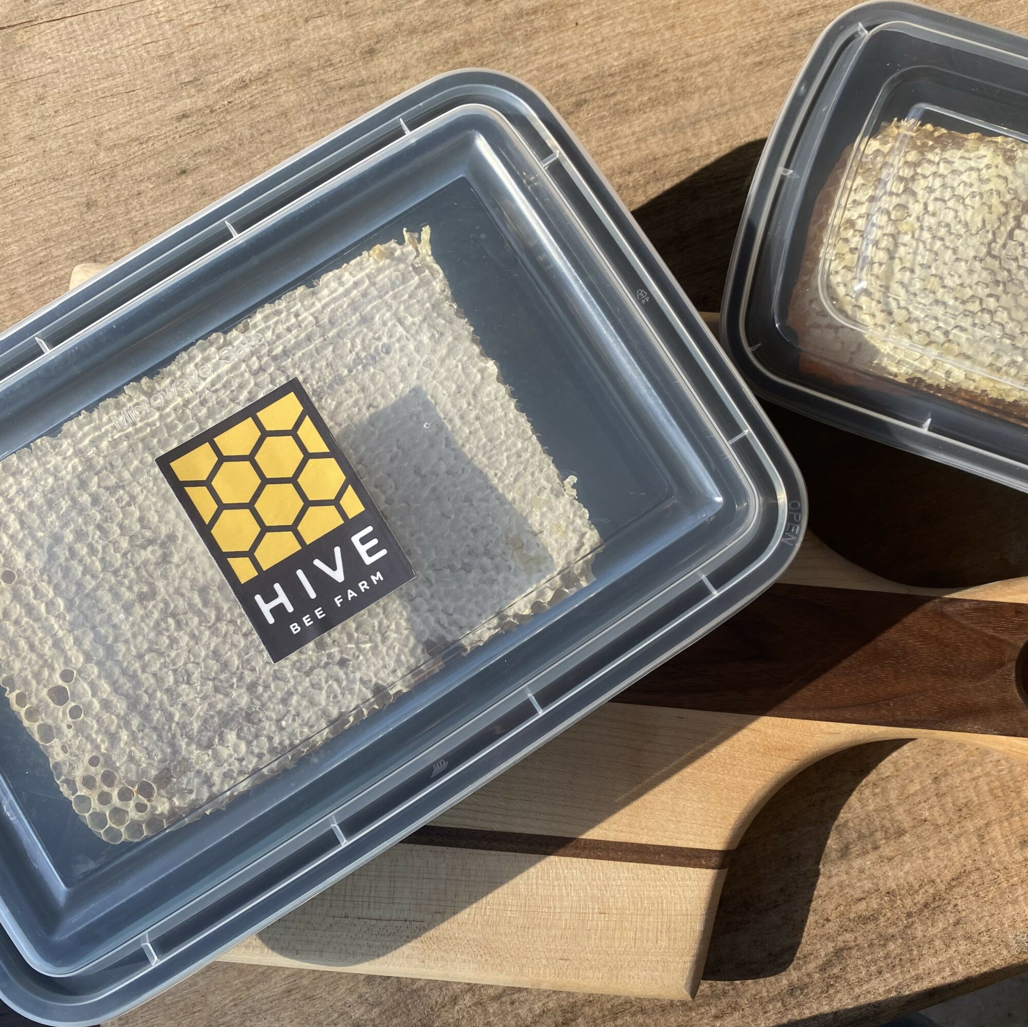 Two plastic containers with honeycomb inside, placed on a wooden surface outdoors, sunlight casting shadows.