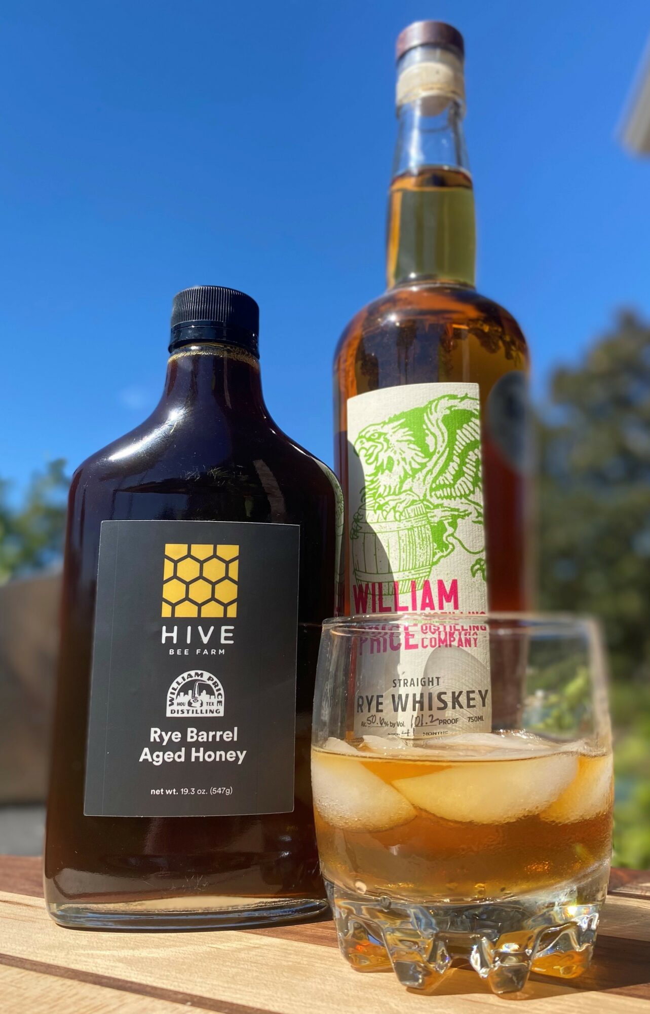 Bottle of Hive Rye Barrel Agave Honey and a glass of whiskey with ice outdoors against a blue sky.