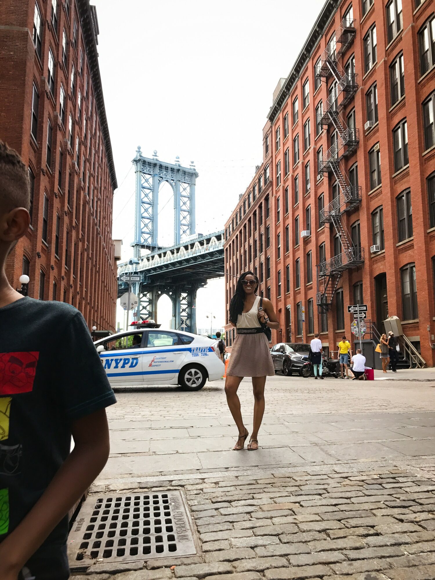 Woman standing in street between red brick buildings with bridge in background, police car nearby, people walking around.