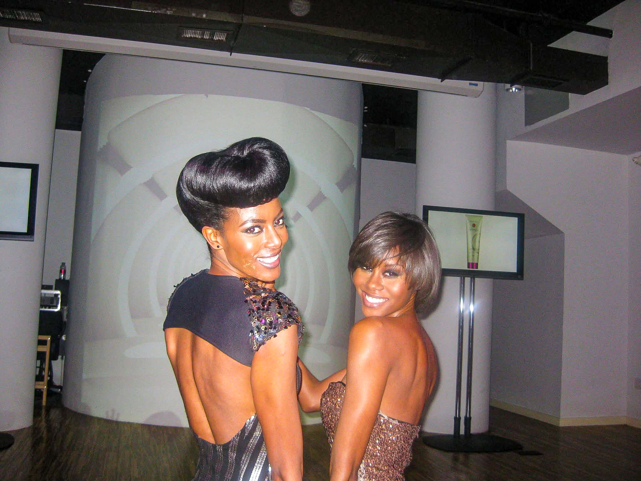 Two women with styled hair smiling, standing close together indoors with screens and a curved wall behind them.