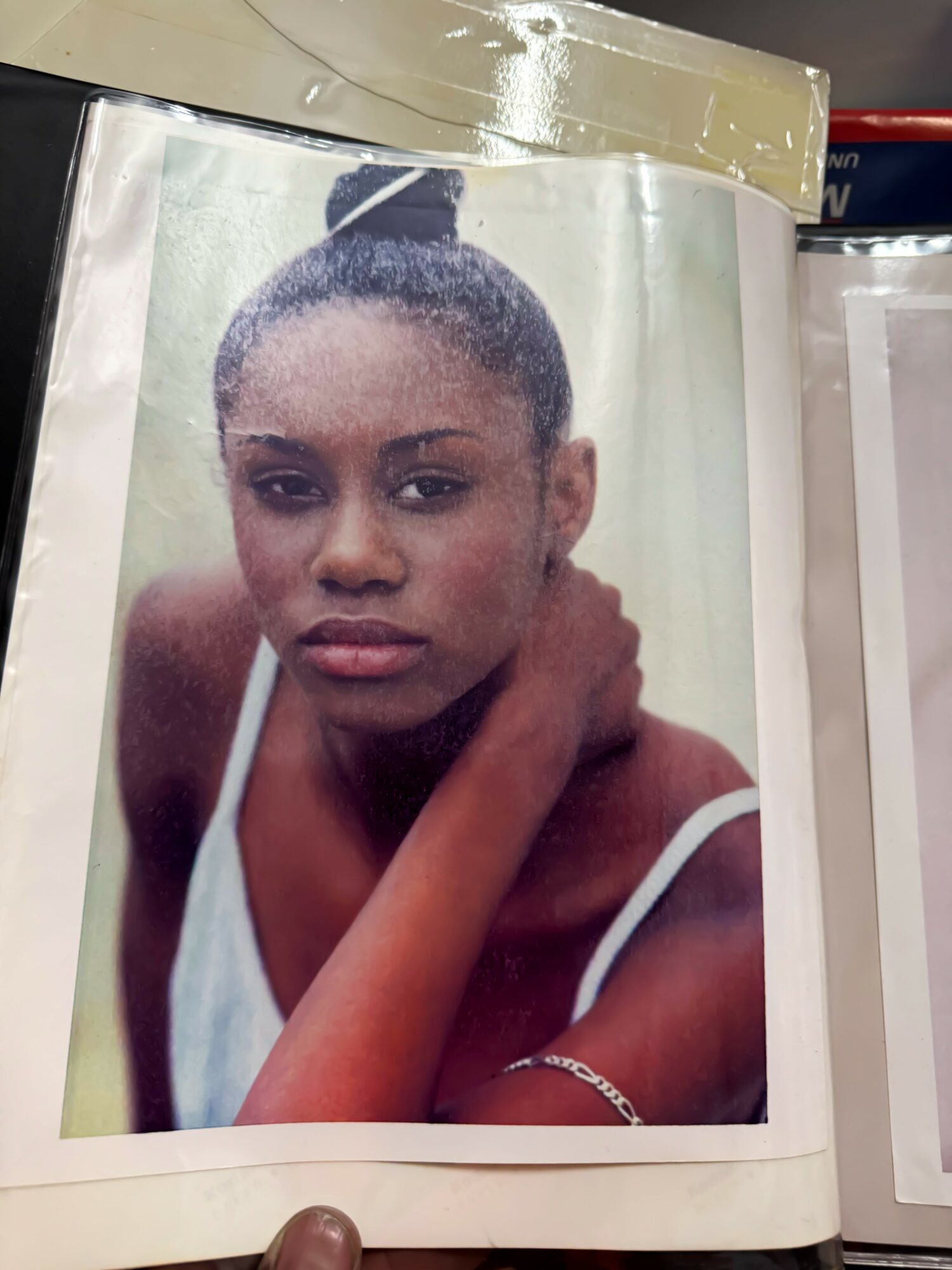 Portrait of a young woman with a serious expression, wearing a white top, with her hand resting on her neck.