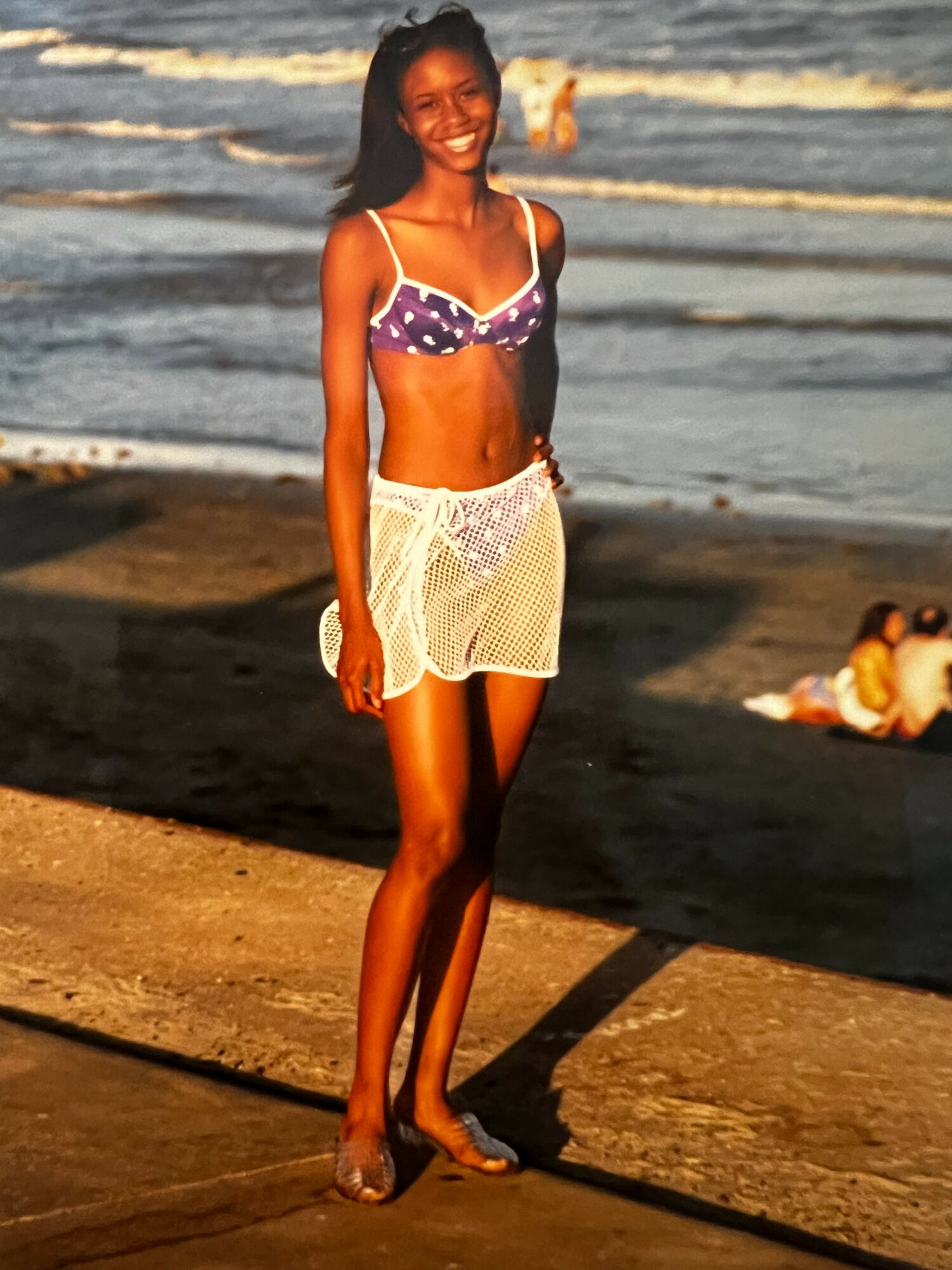 Young woman in a purple bikini top and white skirt standing on a beach with ocean waves in background.