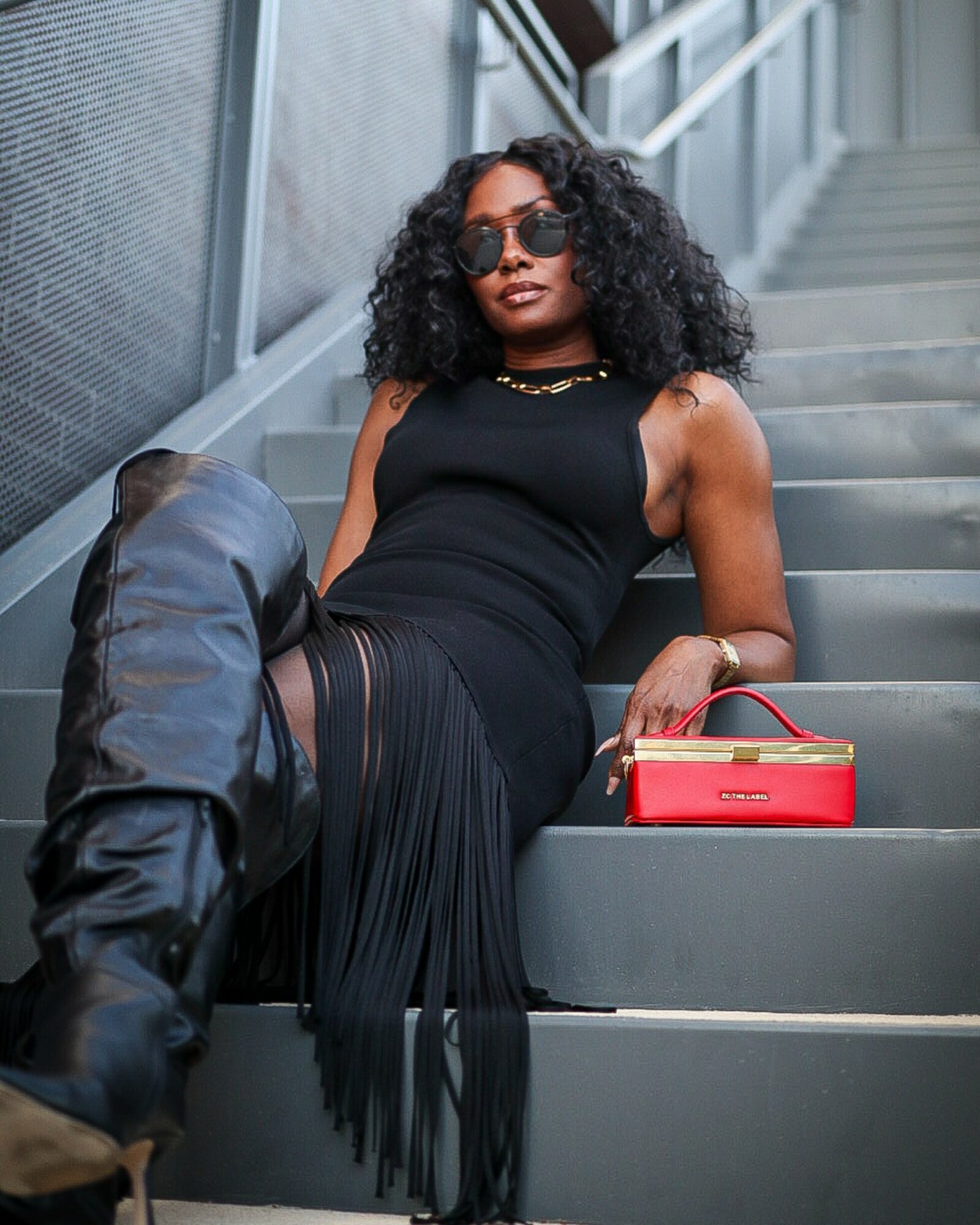 Woman with curly hair wearing sunglasses, black dress, sitting on stairs with a red purse and a black bag.