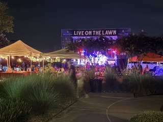 Night scene of an outdoor concert with tents, trees, and a stage with colorful lights, and a sign reading 'LIVE ON THE LAWN'.
