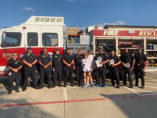 Group of firefighters in uniform standing in front of fire truck, facing camera, outdoors on pavement, clear sky.