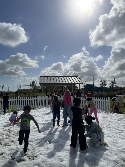 Children and adults playing on snow near a fence, with cloudy sky and sunlight, some children holding snowballs.