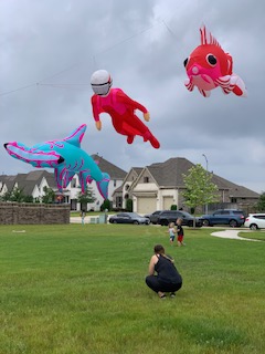 Person kneeling on grass with colorful balloons shaped like animals and characters floating in the sky, in a park setting.