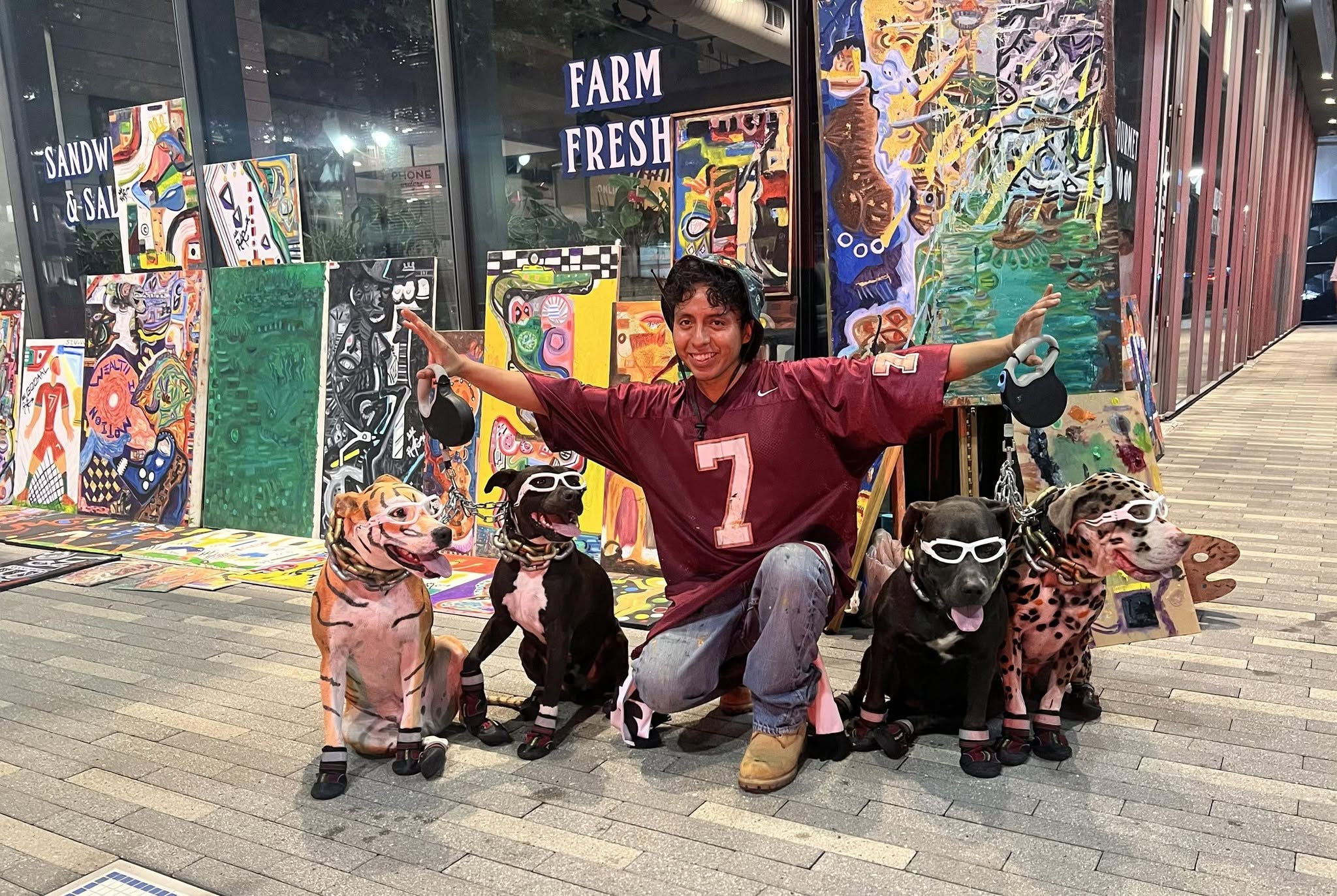 Person in a maroon jersey with number 7 kneels with four dinosaur puppets in front of colorful artwork and windows.