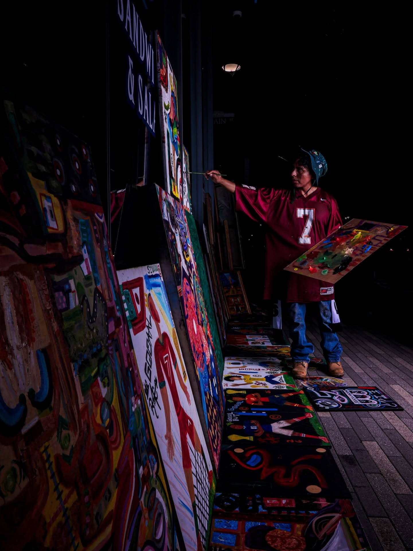 Child in a red jersey painting on a canvas at an art display, with colorful artwork on easels and floor.