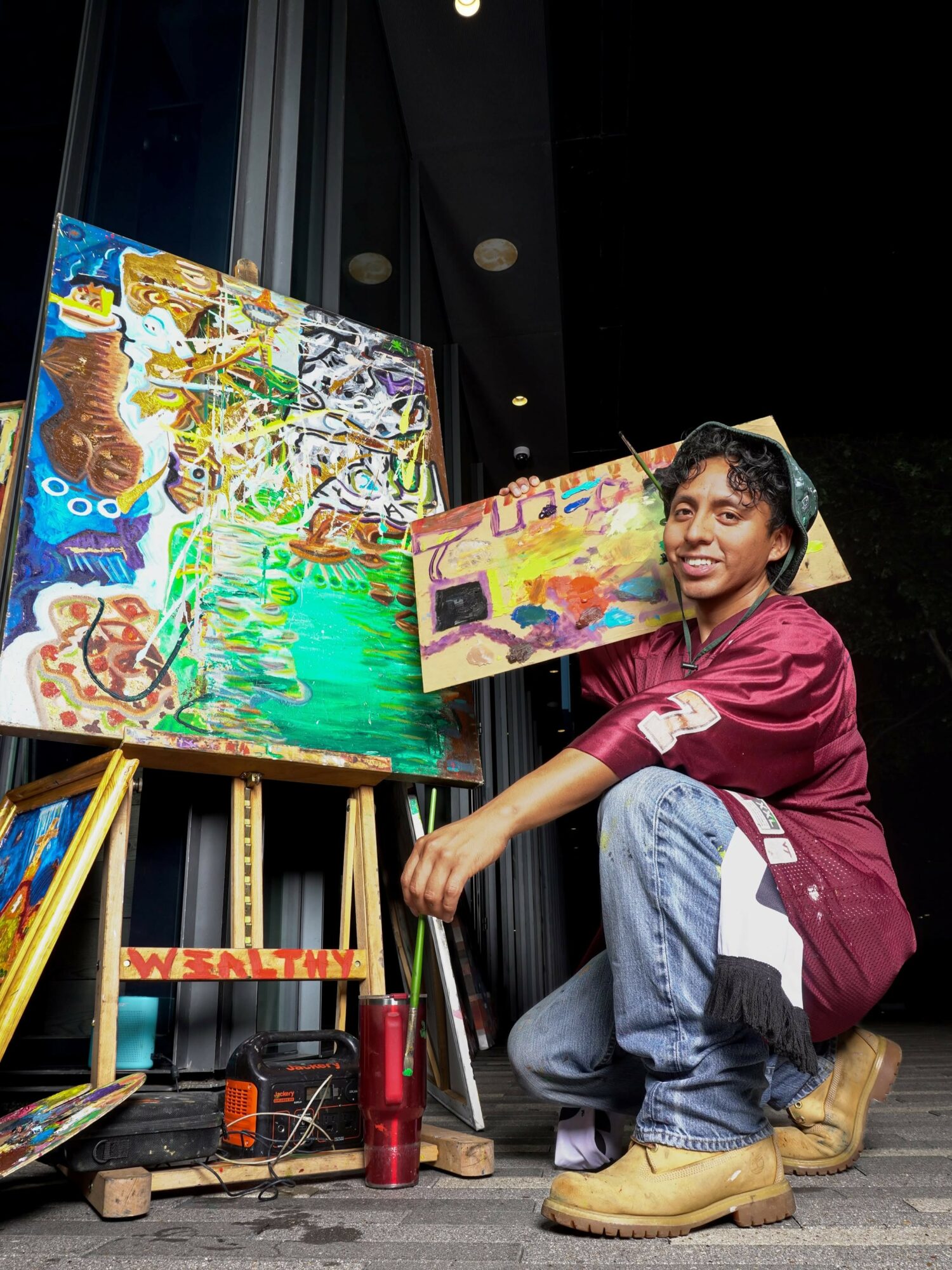 Young person crouching next to colorful paintings on display, wearing a maroon shirt, jeans, and tan boots, indoors.