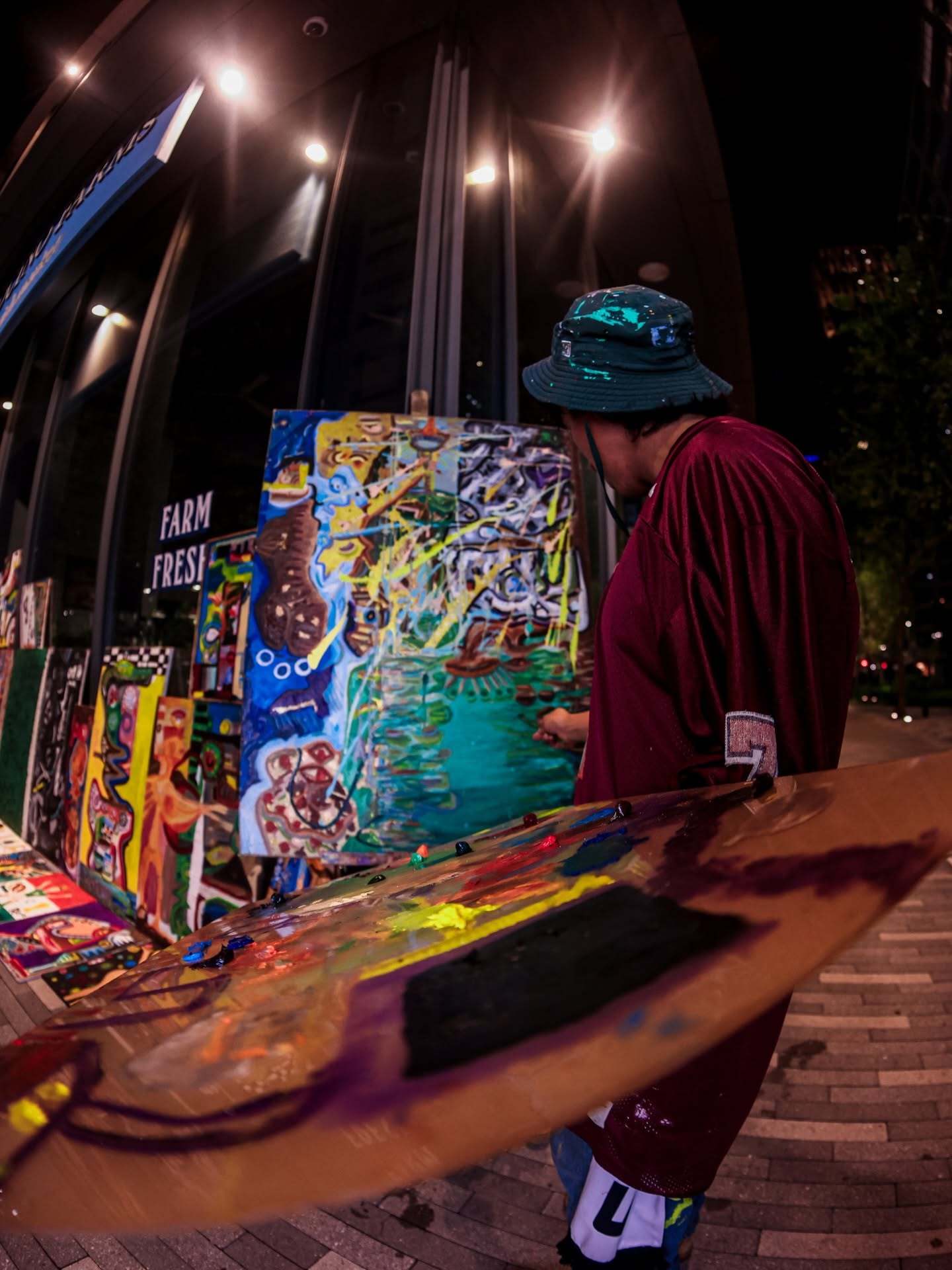 Person wearing a hat and maroon clothing looks at colorful paintings on display at night, with bright lights overhead.